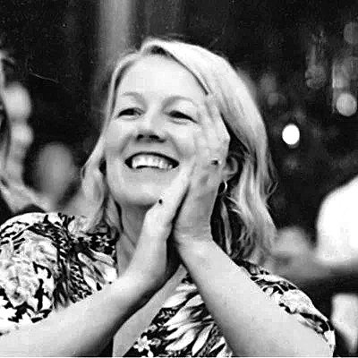 A woman with blonde hair smiling, holding her face with both hands, and wearing a patterned top in an indoor setting.