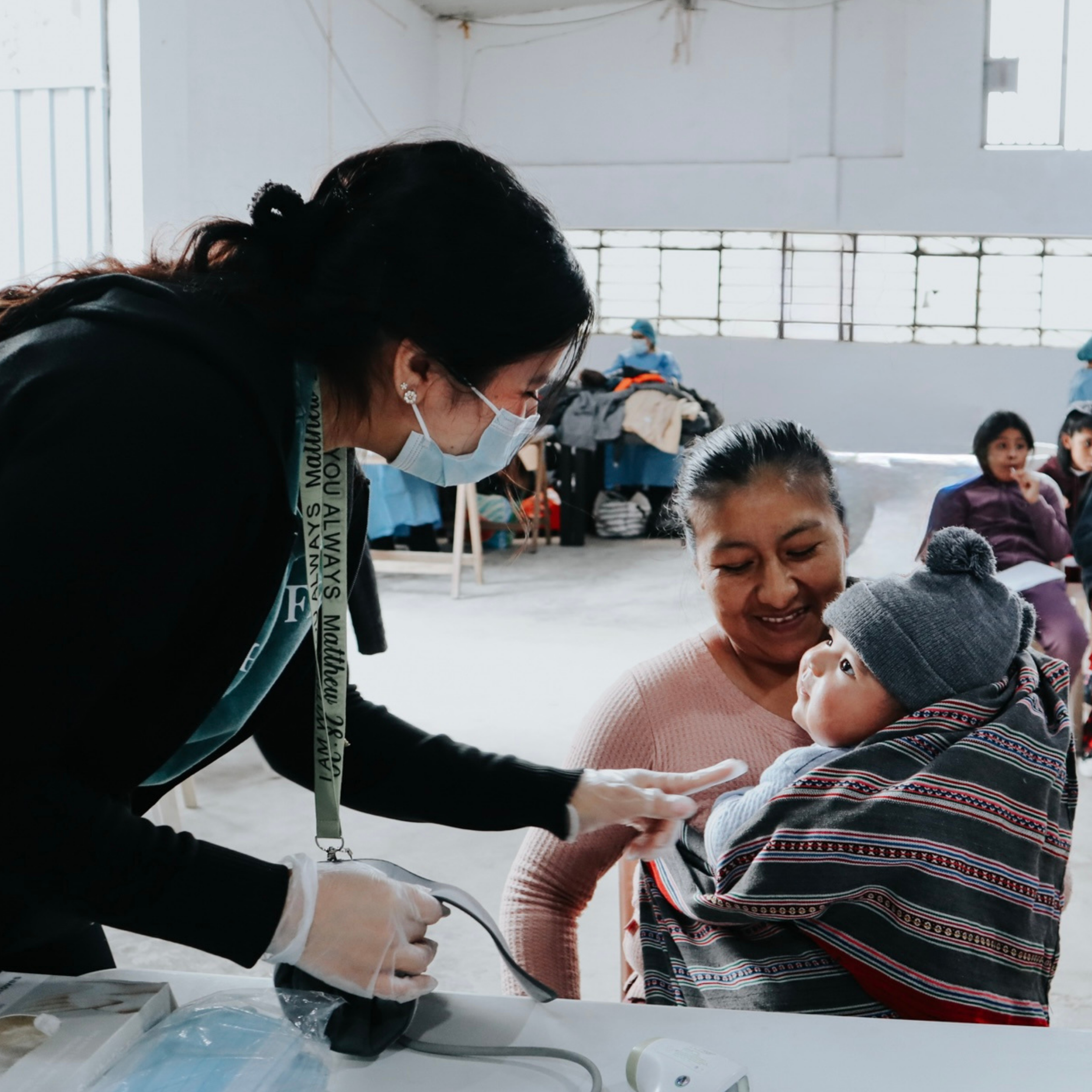 A healthcare worker wearing a face mask, gloves, and scrubs, assisting a young child in a clinical or vaccination setting with medical supplies and equipment around.