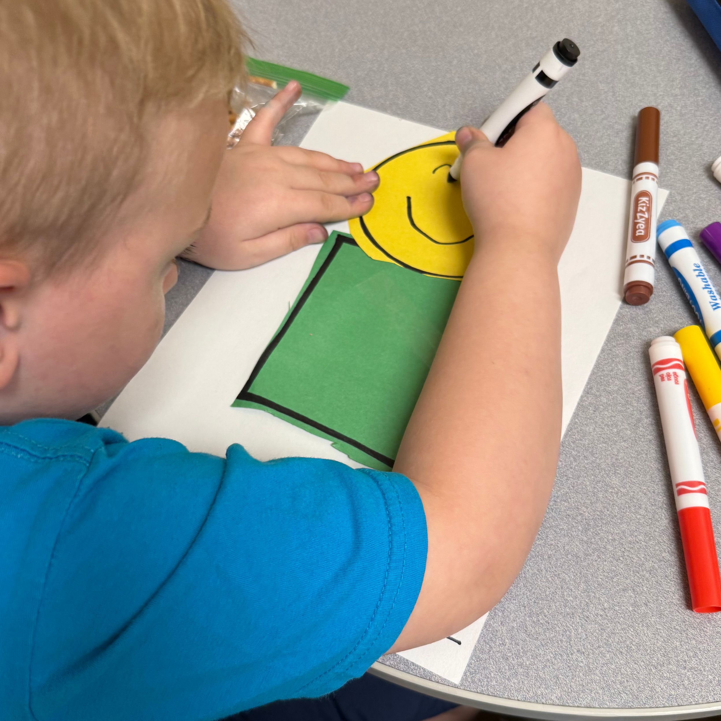 Close-up of a child's hands coloring a yellow smiley face on paper with black marker, with markers and art supplies on a gray table surface.
