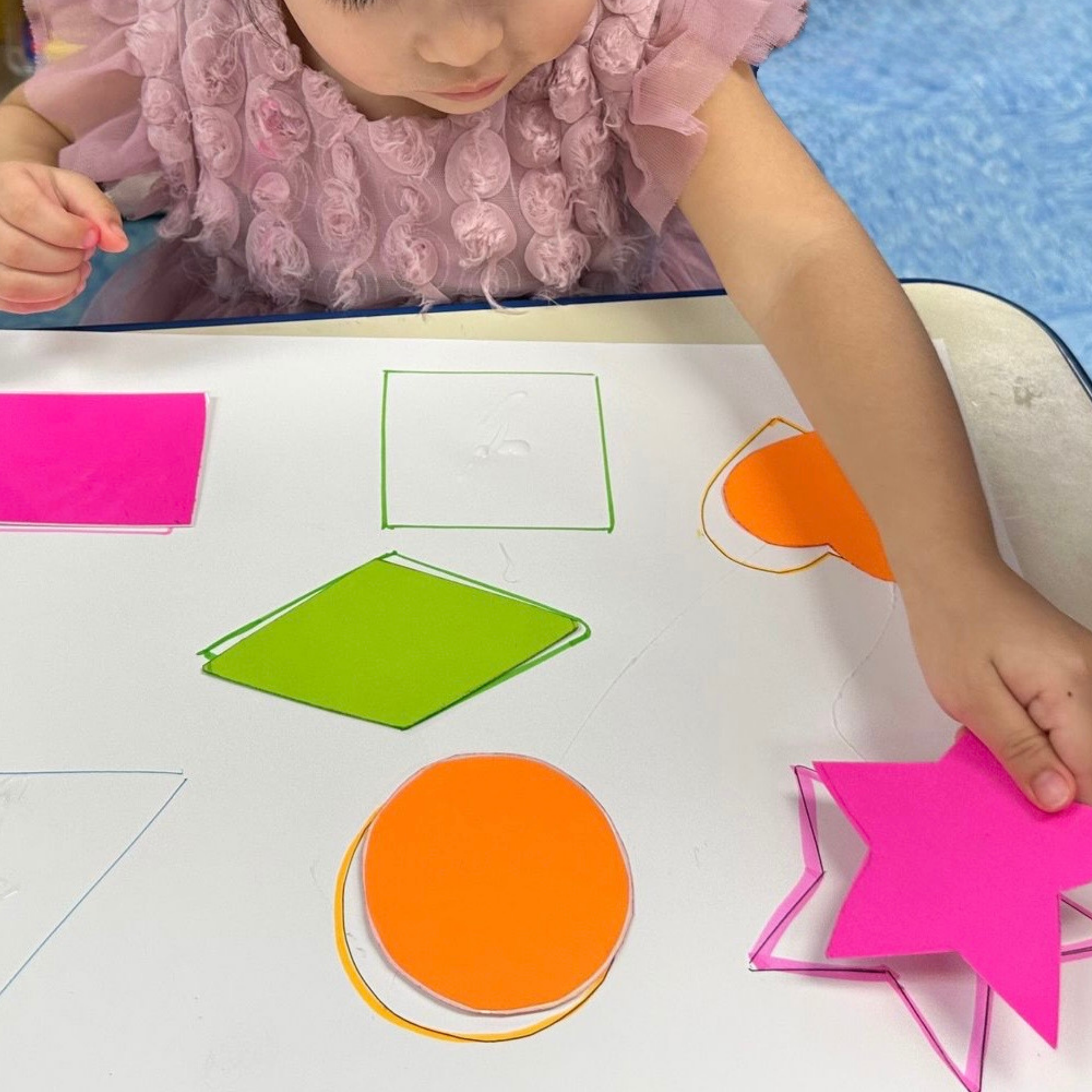 Close-up of a young girl wearing a pink dress with frilled details, coloring on a worksheet with colored paper and paper cutouts on a white table.