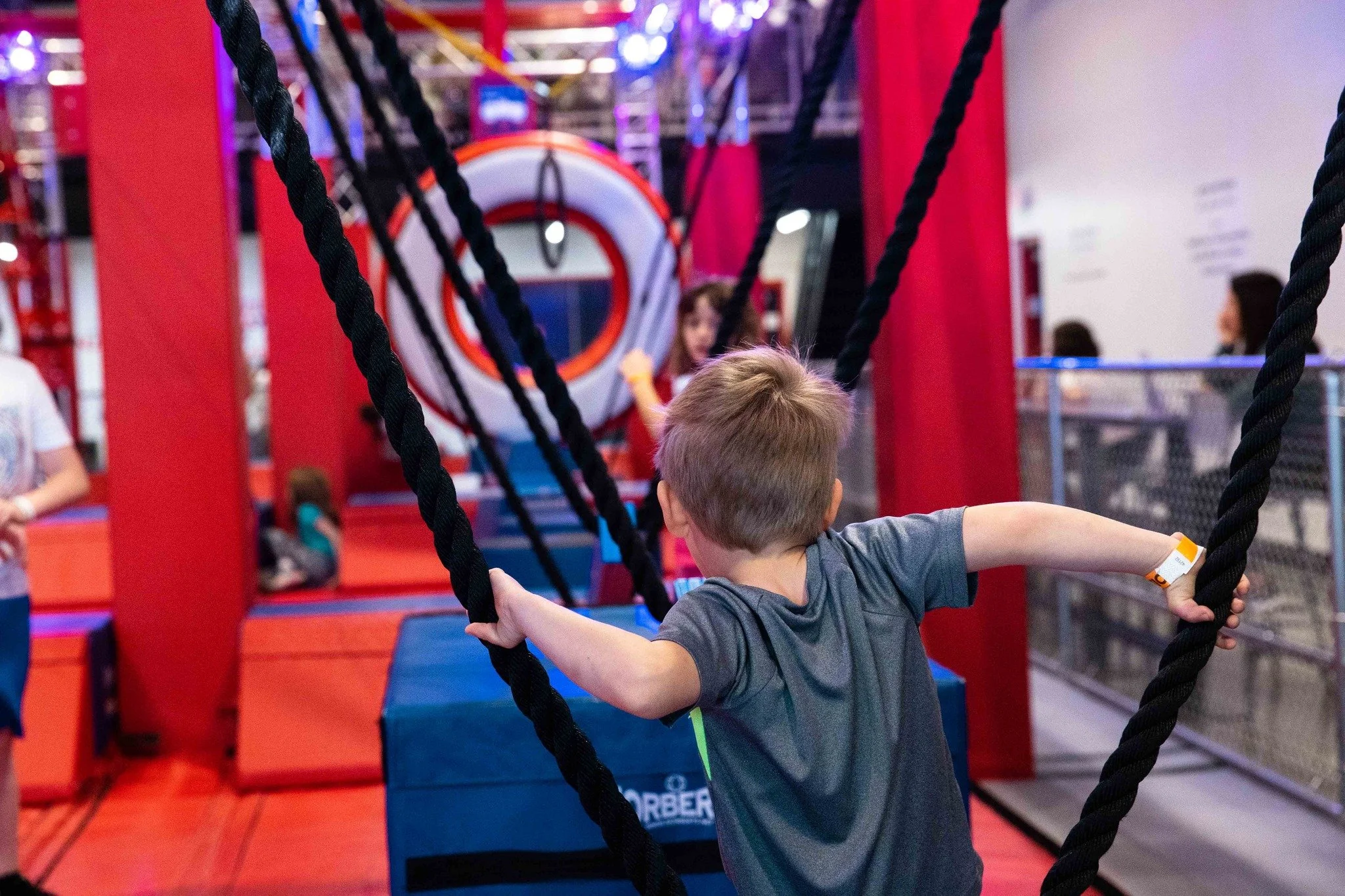 A young boy is playing on a ropes course at an indoor trampoline park. He is holding onto black rope obstacles, with other children and adults in the background, and bright colorful play structures and a mural on the wall.
