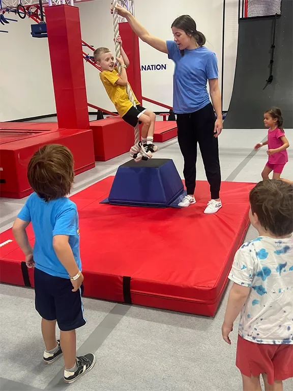 A young boy in a yellow shirt and black shorts is climbing a rope swing while an instructor in a blue shirt assists him. Three other children are watching nearby inside an indoor playground with red mats, padded walls, and climbing equipment.