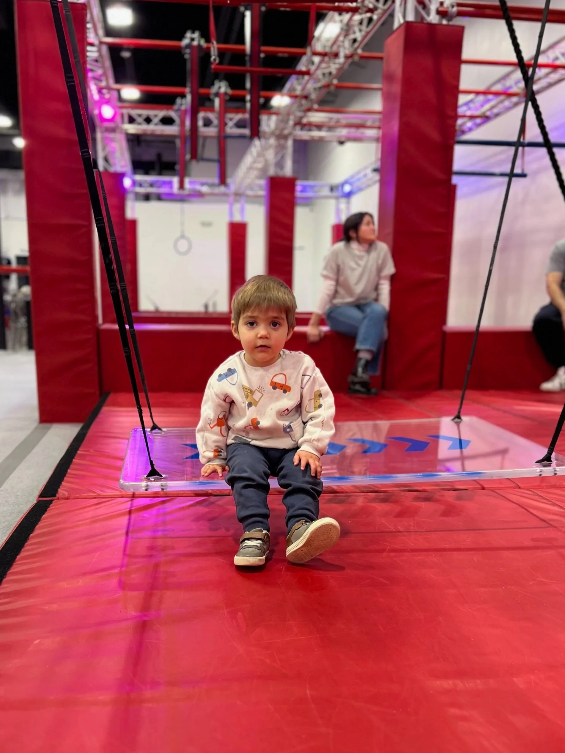A young boy sitting on a trampoline with red padding, with a woman sitting on a red structure in the background. The trampoline is part of a larger indoor play area with metal and plastic framework, purple and pink lighting, and other children in the background.