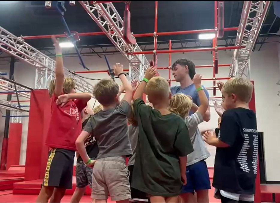 A group of kids gathered around their trainer at an indoor ninja obstacle course, listening and engaging during a training session.
