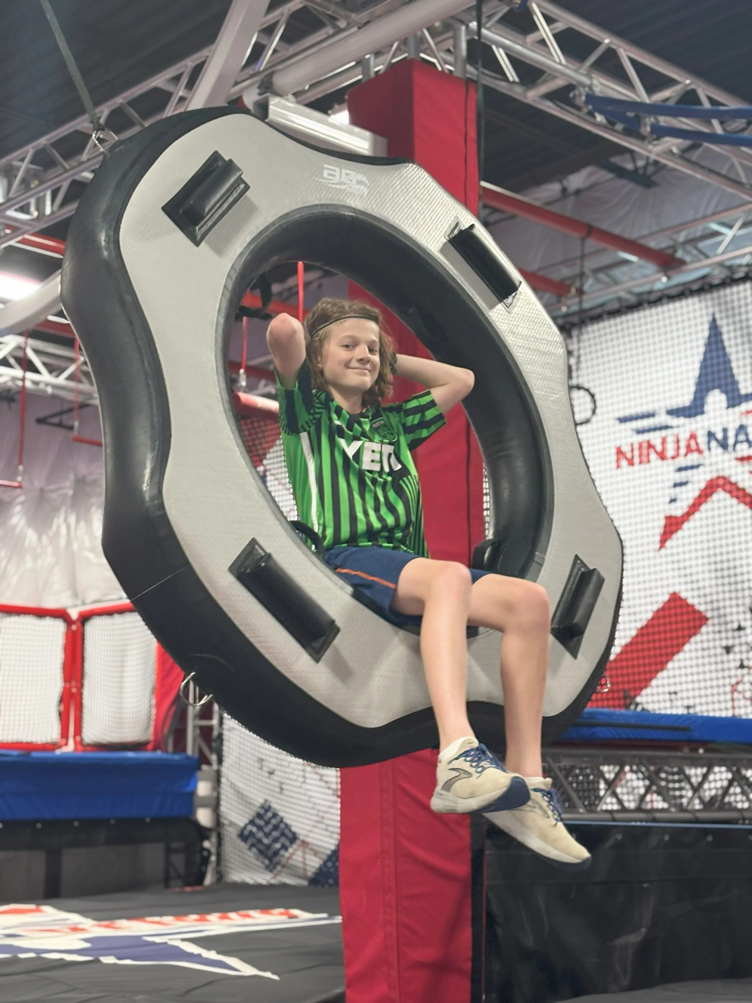 A young boy with curly hair sitting in a circular amusement park ride or game on a trampoline-like structure, wearing a green and black striped sports jersey and sneakers, with his hands behind his head and smiling.