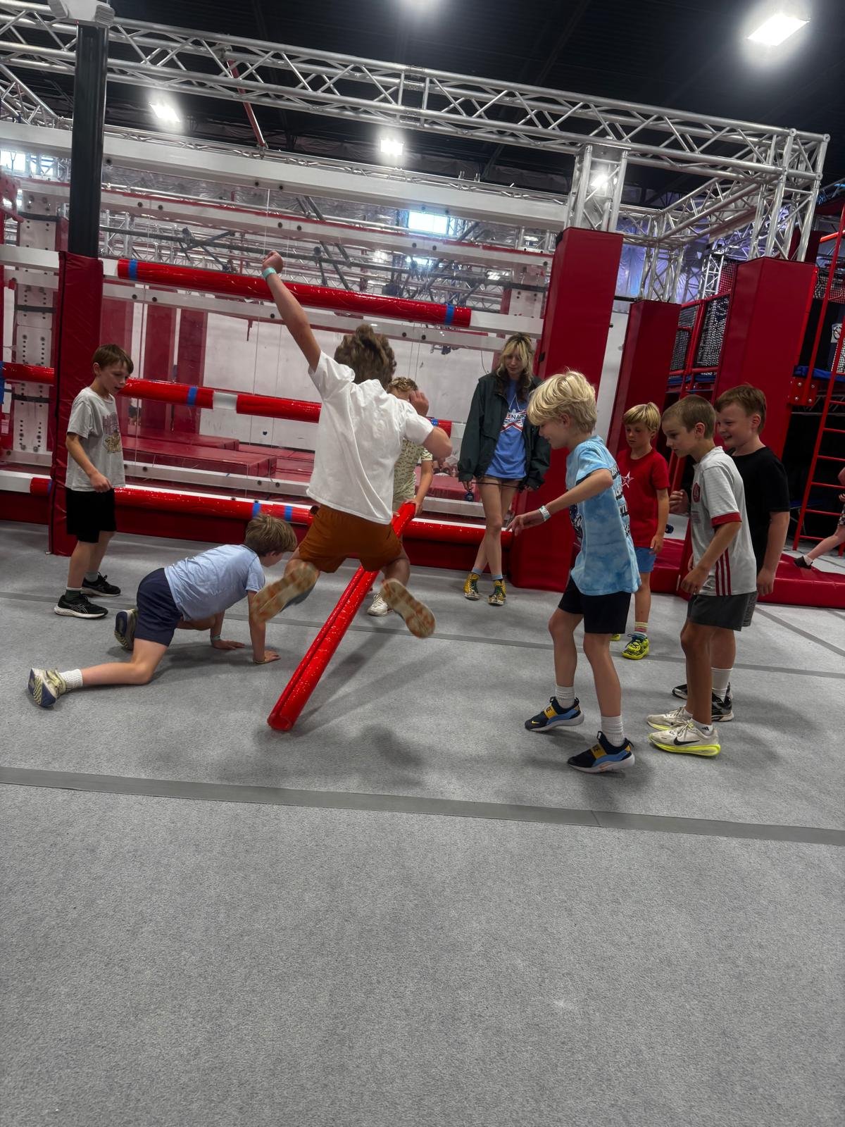 Children playing on an indoor adventure playground with red and grey obstacle course, surrounded by safety mats.