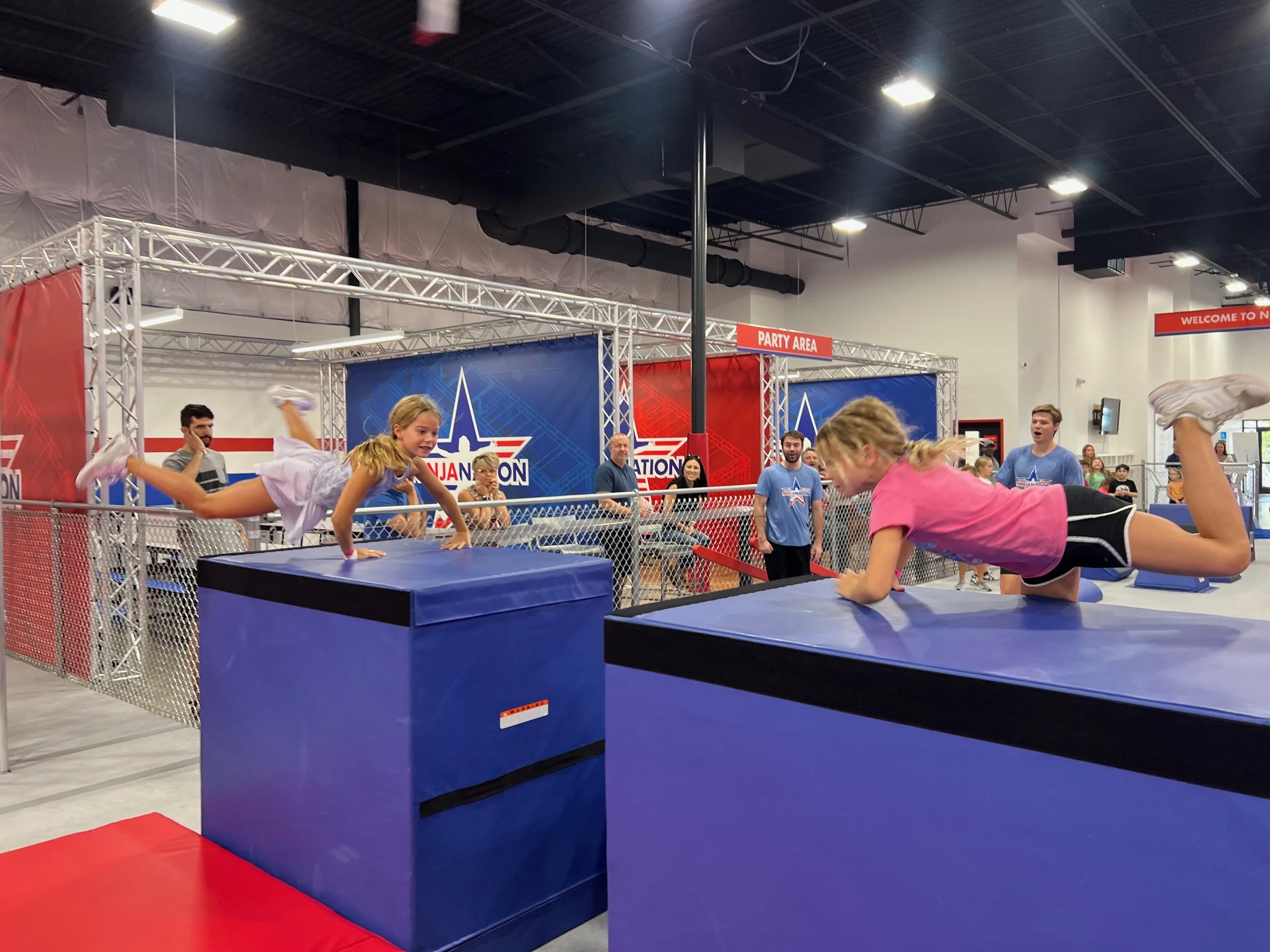 Children and adults at an indoor ninja obstacle course, with a girl doing a handspring over a foam block and others watching.
