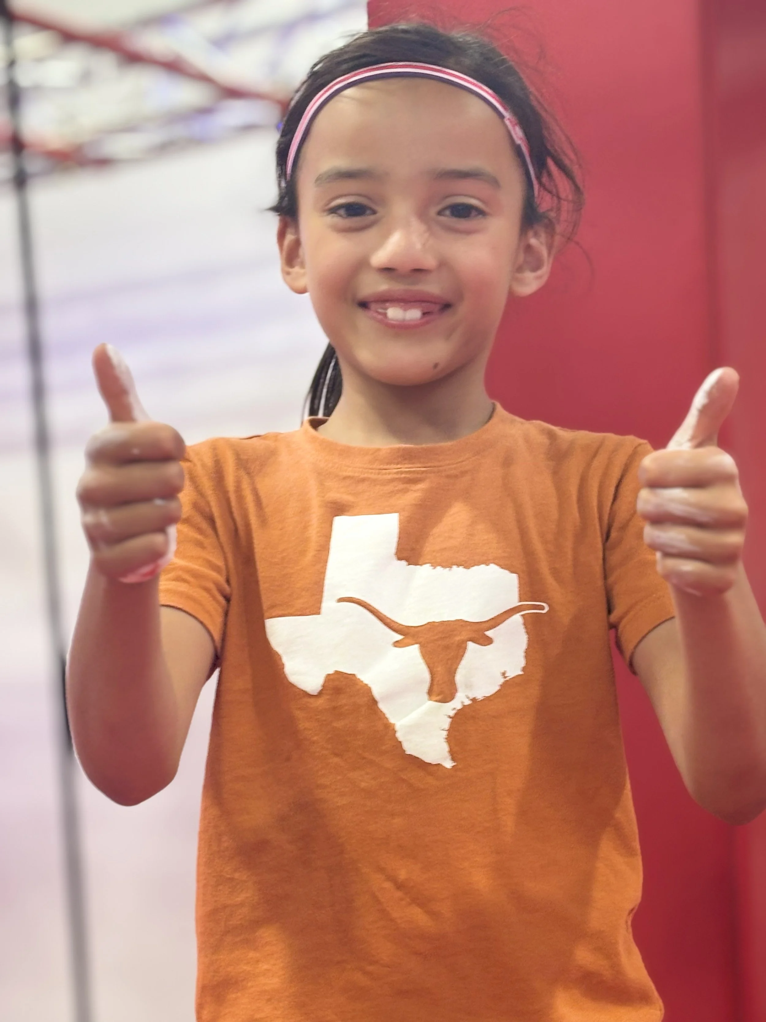 A young girl with dark hair wearing a pink headband and an orange Texas Longhorns t-shirt, giving two thumbs up inside a building with a red wall behind her.