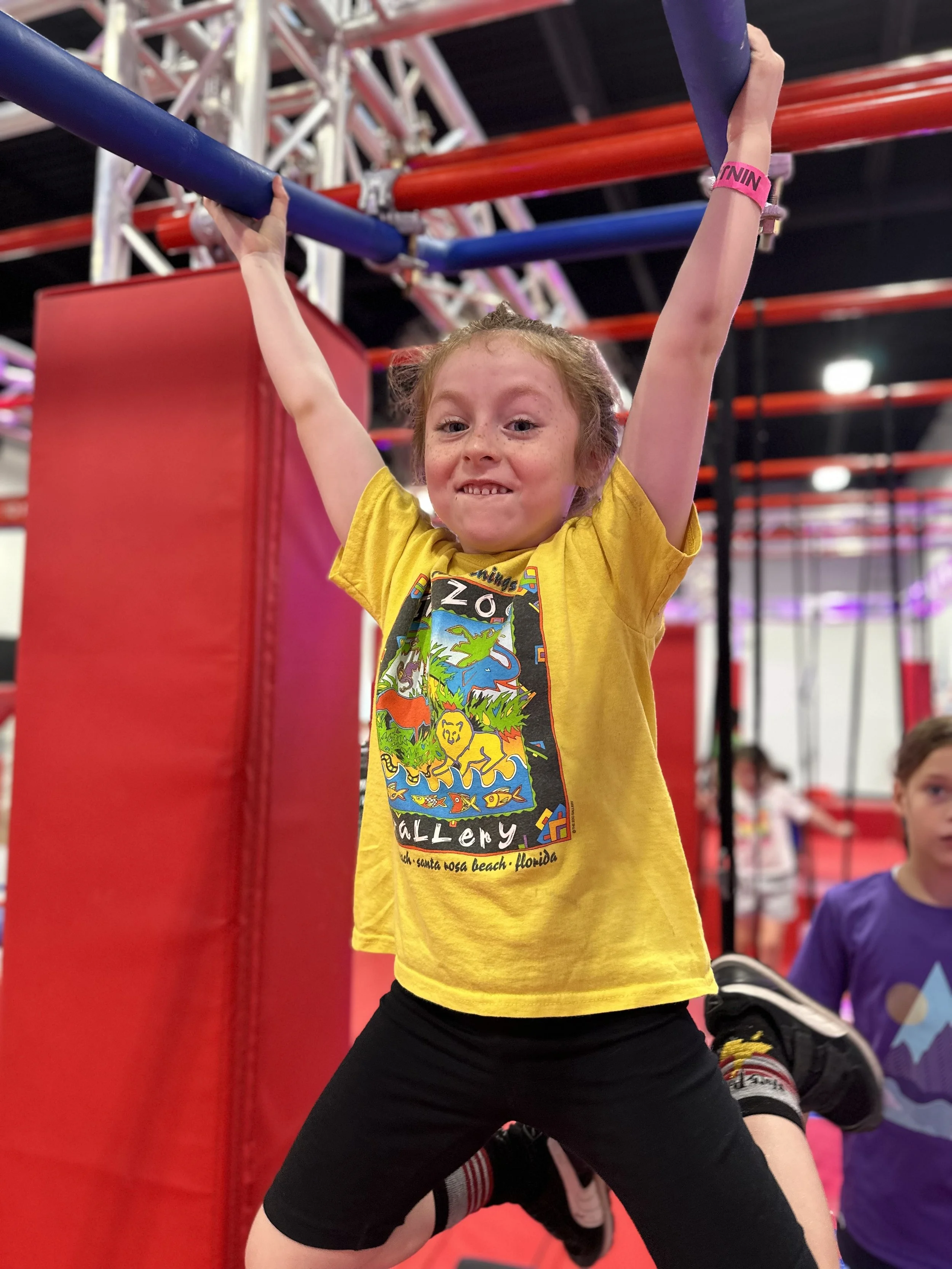 Young girl in a yellow t-shirt with a colorful dinosaur design, hanging from gymnastic bars in an indoor playground or gym, with other children nearby.
