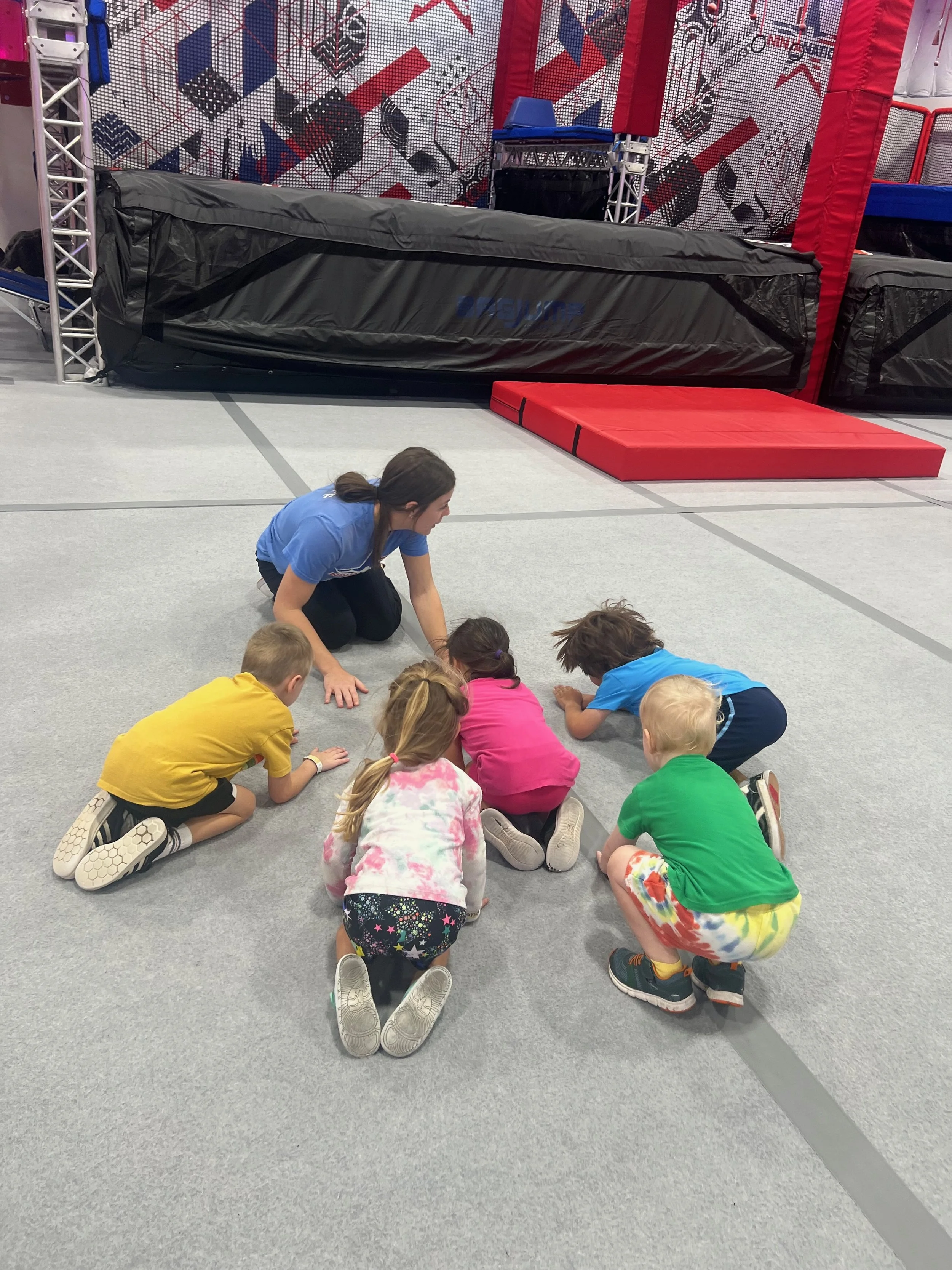A group of children and a woman with dark hair tied back, kneeling on the floor, all facing down and engaged in an activity, inside a gym with sports mats and padded walls.
