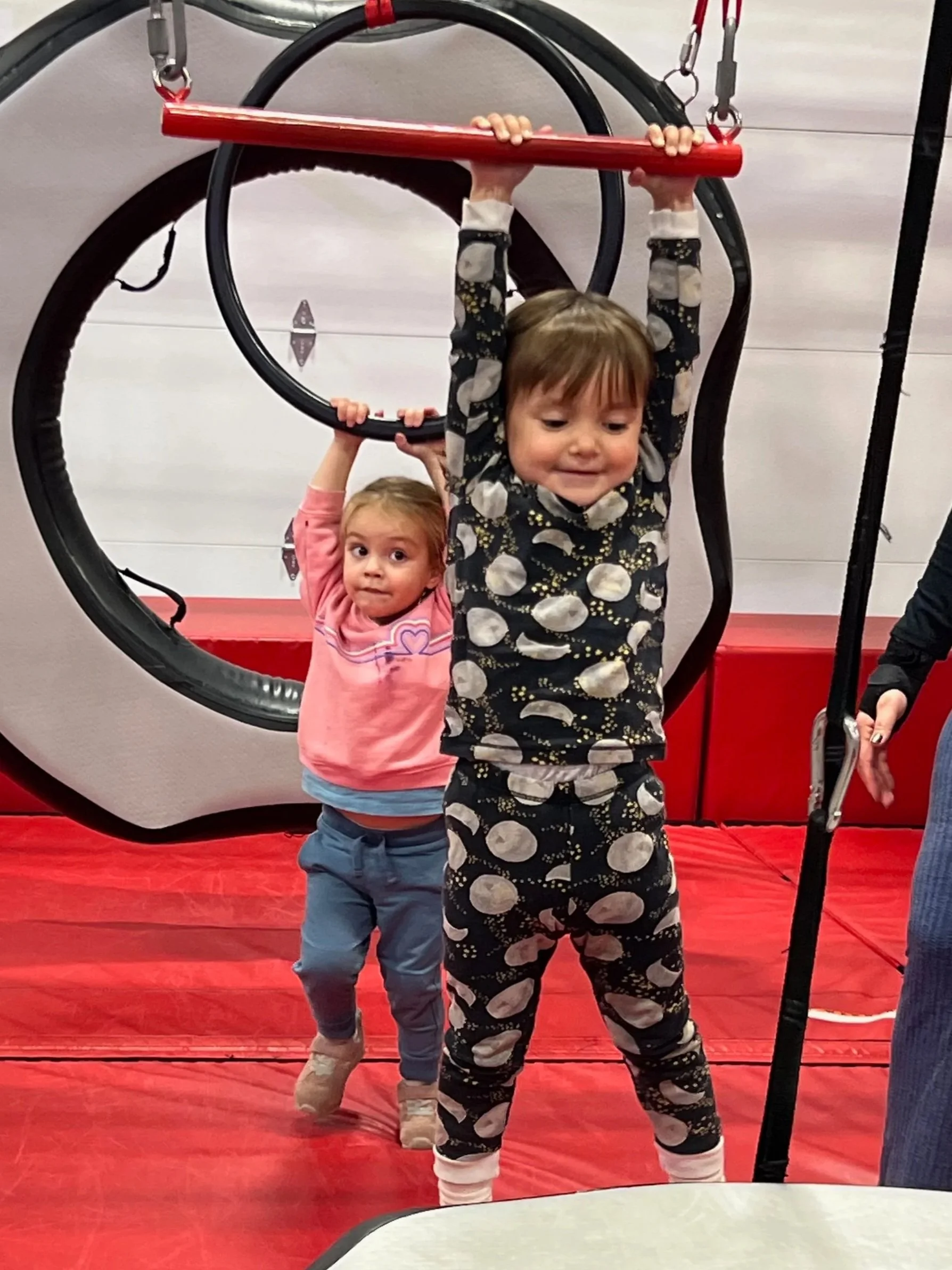 Two young children playing on a circus-themed climbing frame with red, black, and white elements. The background has a gray wall, and the floor is covered with a red mat.