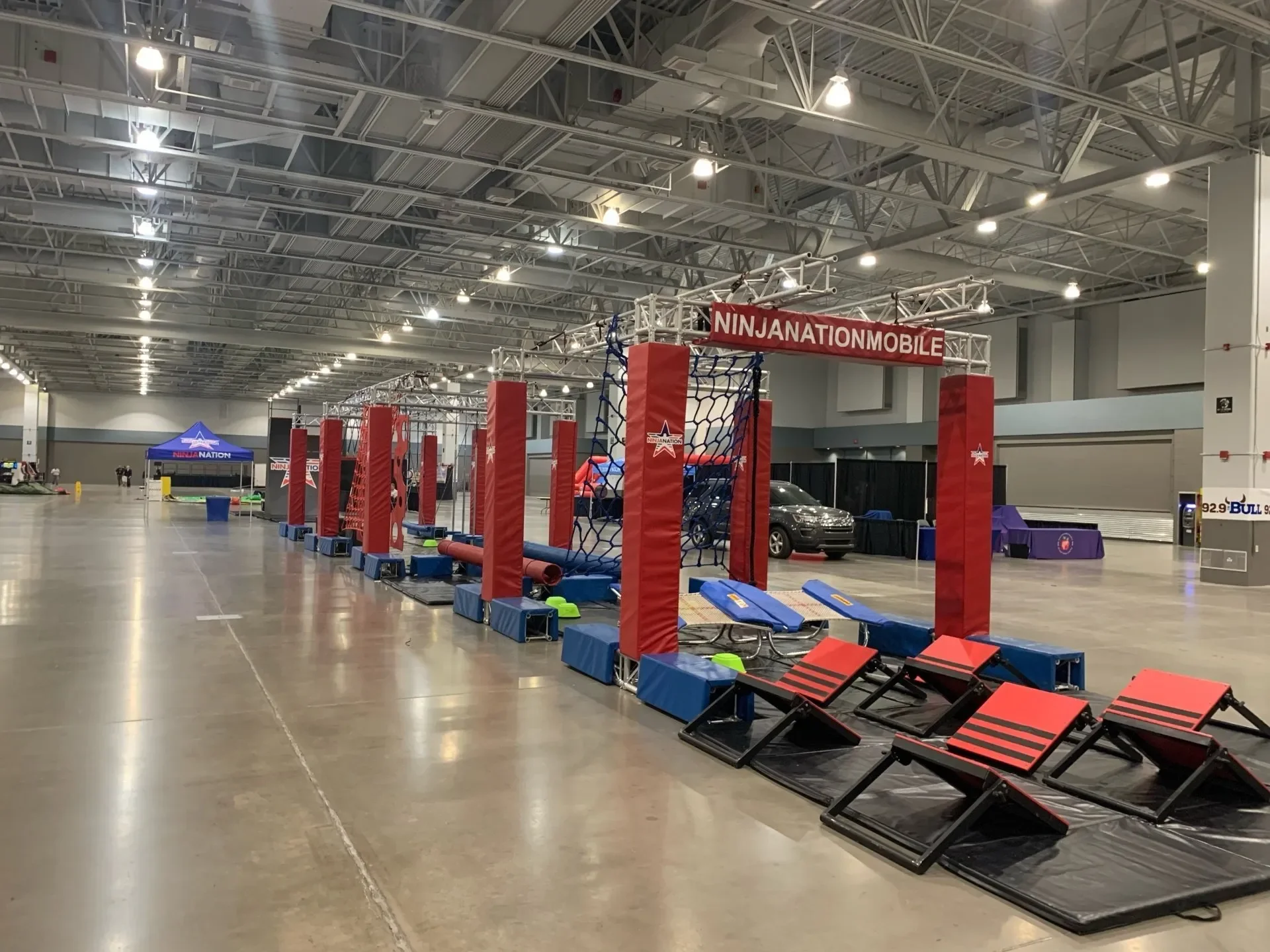Indoor Ninja Warrior obstacle course setup with padded mats, climbing nets, and ramp challenges. A blue tent with red and white branding is visible in the background, along with a parked car and some equipment.