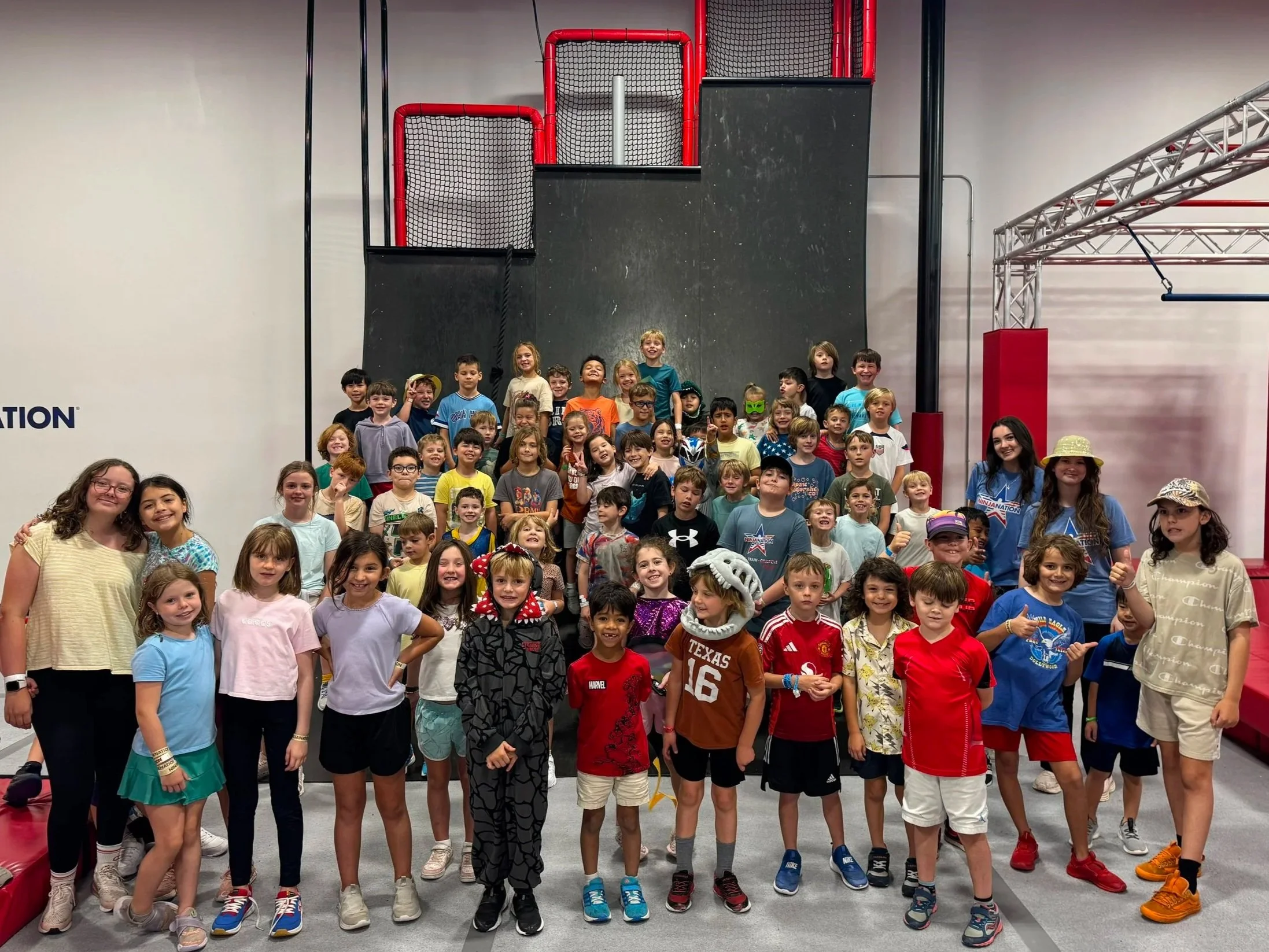 Group of children and adults posing together inside a gymnasium or indoor play area. The children are smiling and wearing casual clothes and some have costumes. The background features black and red padded walls, a climbing net, and metal gym equipment.