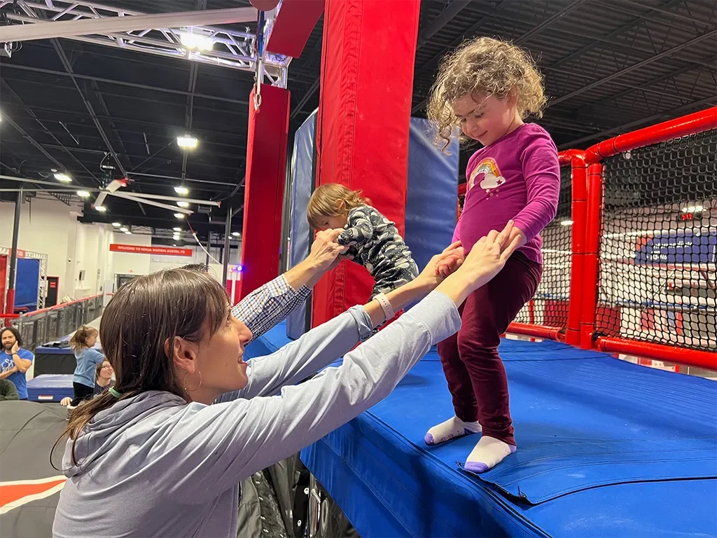 A woman helping a young girl walk on a trampoline at an indoor play area. Another child is playing in the background.