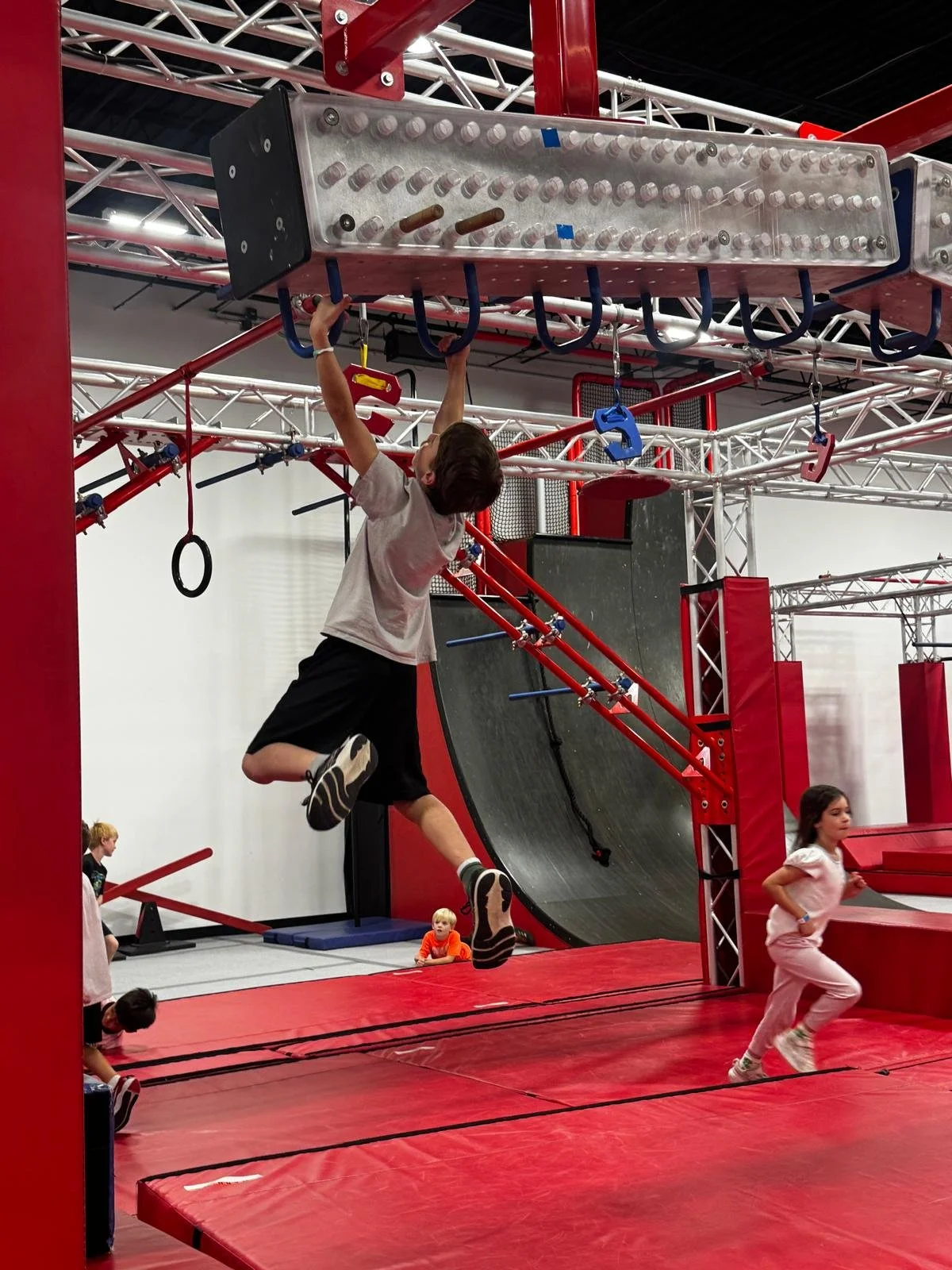 Young boy climbing a blue metal structure at an indoor ninja obstacle course, with other children around and a large black ramp in the background.