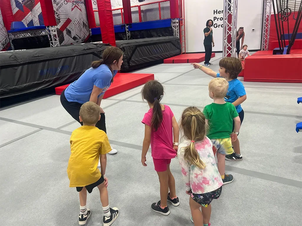 Children at a trampoline park with an instructor, some children are waiting while one child points and talks, colorful padded inflatables in background.
