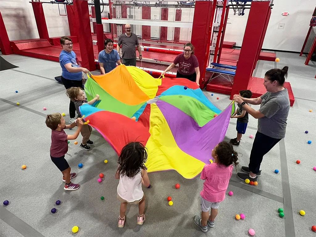 Children and adults playing with a colorful parachute inside a playroom, scattered with small colorful balls.