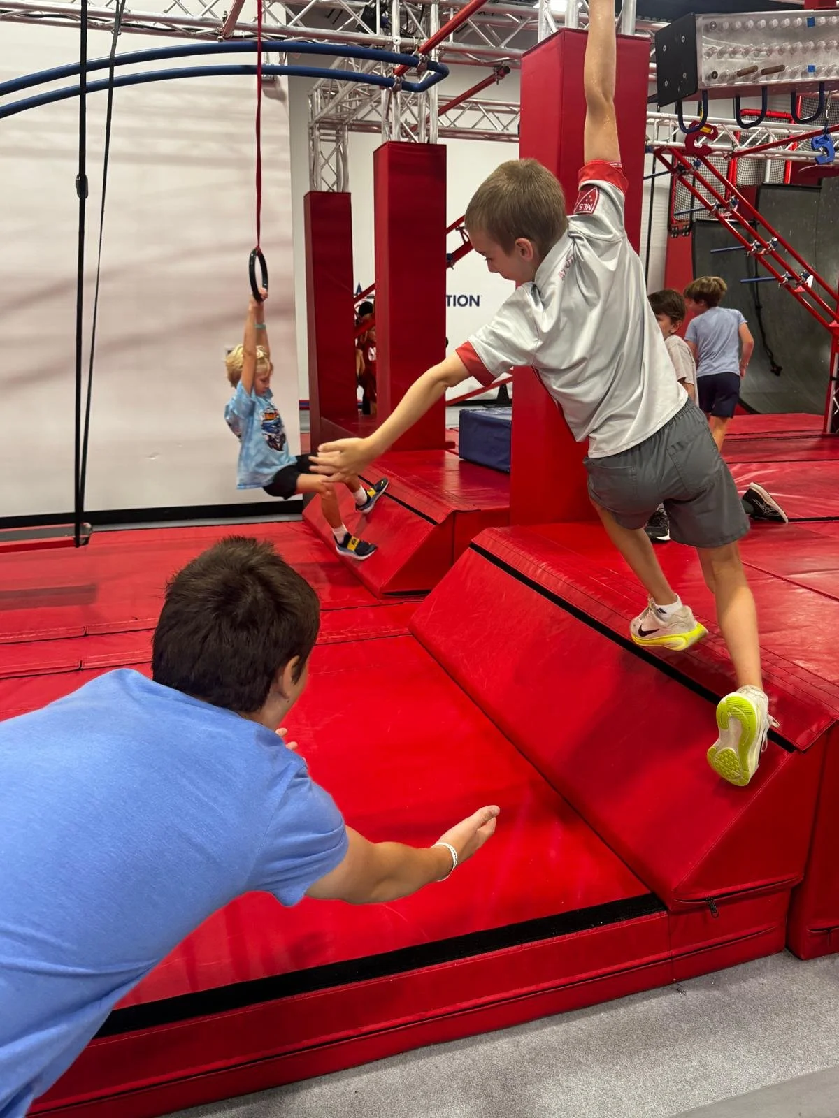 Children playing on a red indoor obstacle course, with one child hanging on gymnastic rings and another helping him climb a padded platform.