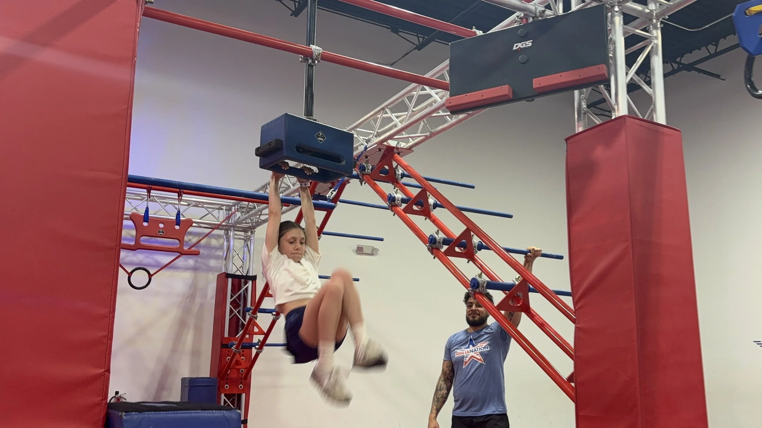 A young girl participating in an obstacle course, hanging from a blue block on a red metal climbing structure at Ninja Nation.