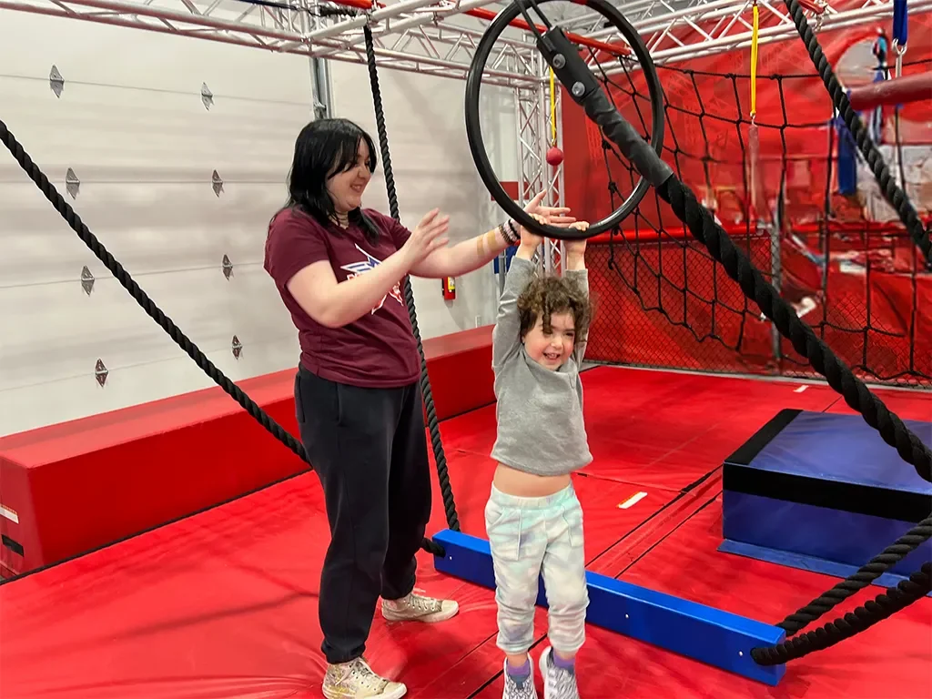 A woman helping a young girl hang from a large circular trap in an indoor play area with red padded flooring and climbing structures.