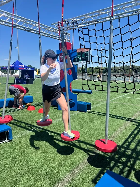Girl wearing athletic clothes and a cap crossing an obstacle course on a grassy field during daytime.