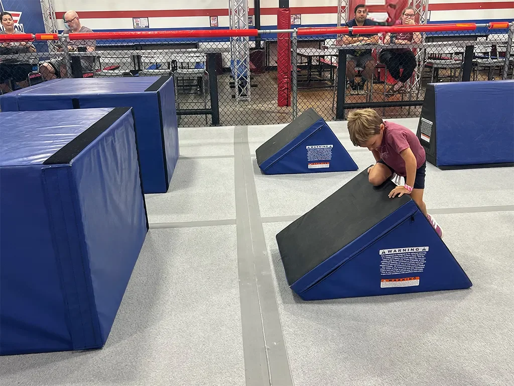 A young boy climbing over blue padded gym mats in an indoor recreational facility, with several adults sitting at tables in the background.