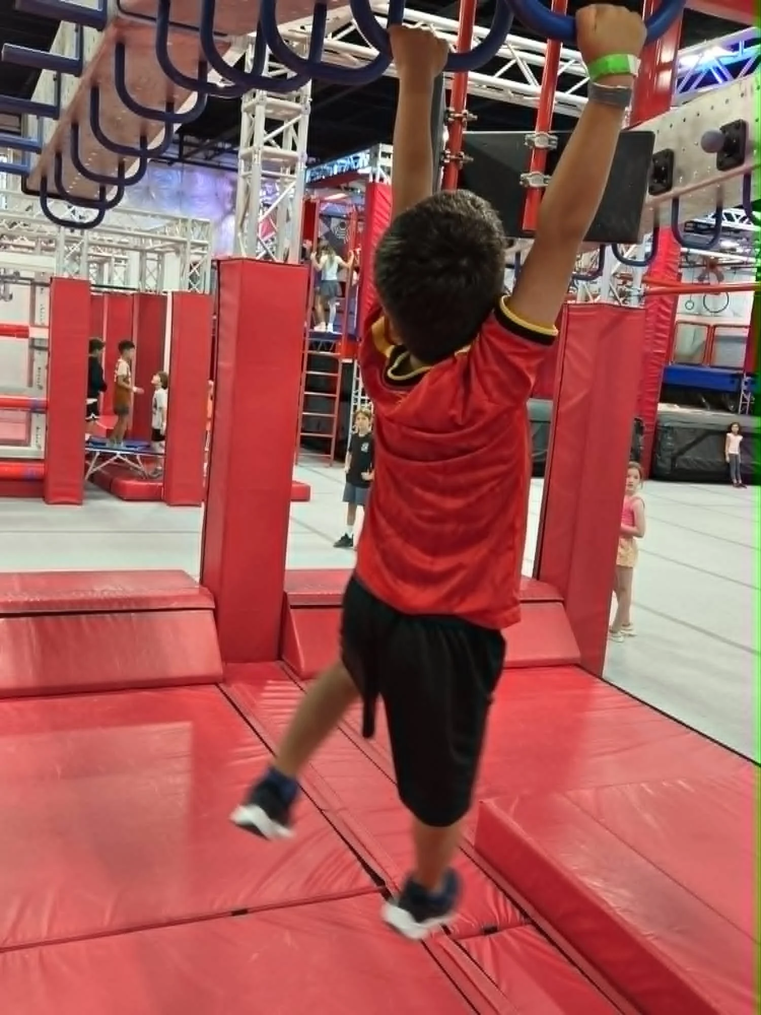 A boy hanging from a blue gymnastic bar at an indoor amusement park or arcade, with other children and adults in the background.