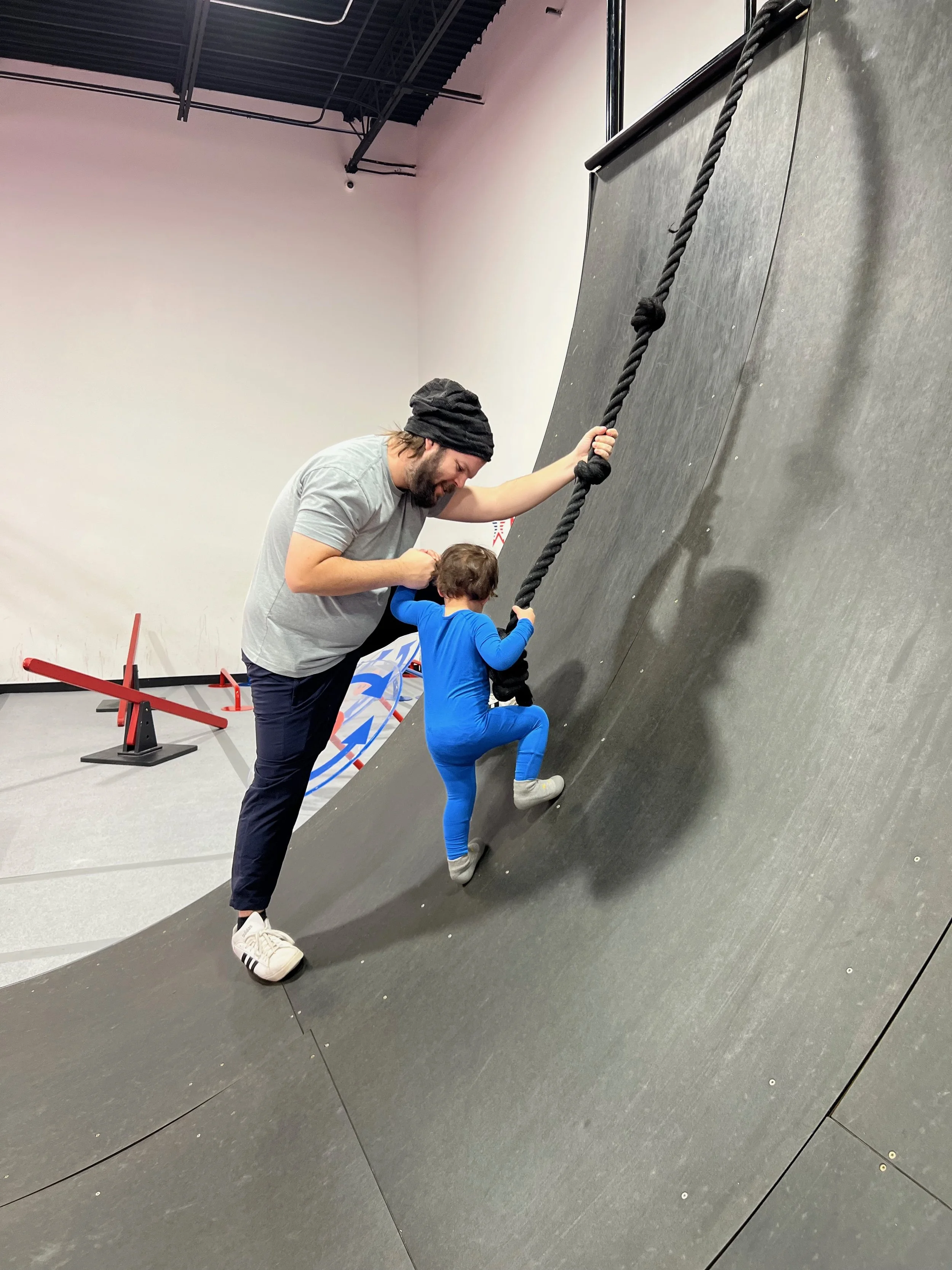 A man and a young girl are at an indoor rock climbing facility. The man is helping the girl with her climbing harness as she prepares to climb a vertical wall with a large rope hanging from the top.