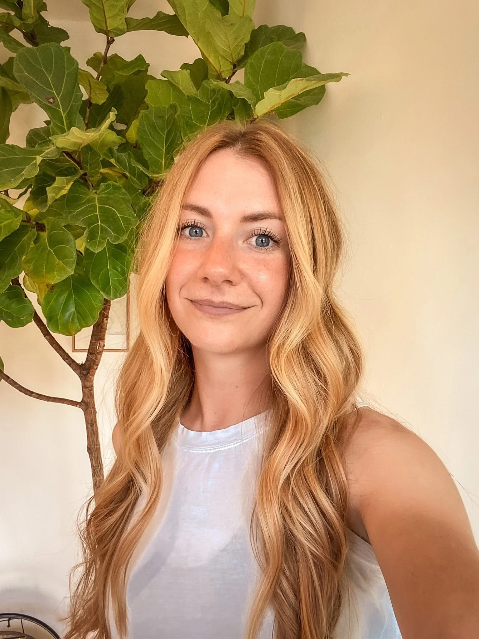 A woman with long, wavy light colored hair and blue eyes, smiling, standing in front of a leafy green plant with large leaves against a plain beige wall.