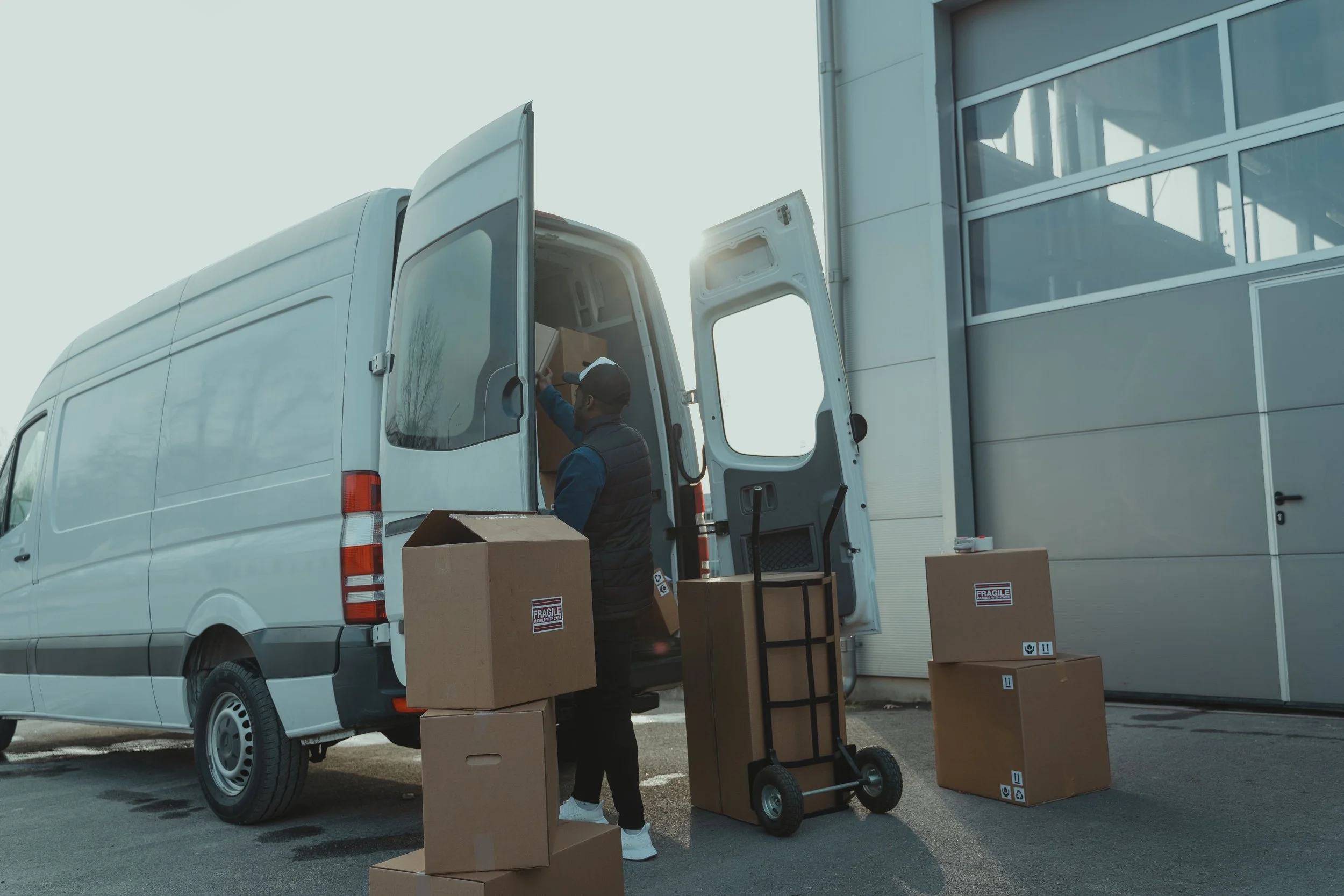 Person loading boxes into the back of a delivery van parked outside a modern building with large glass windows.