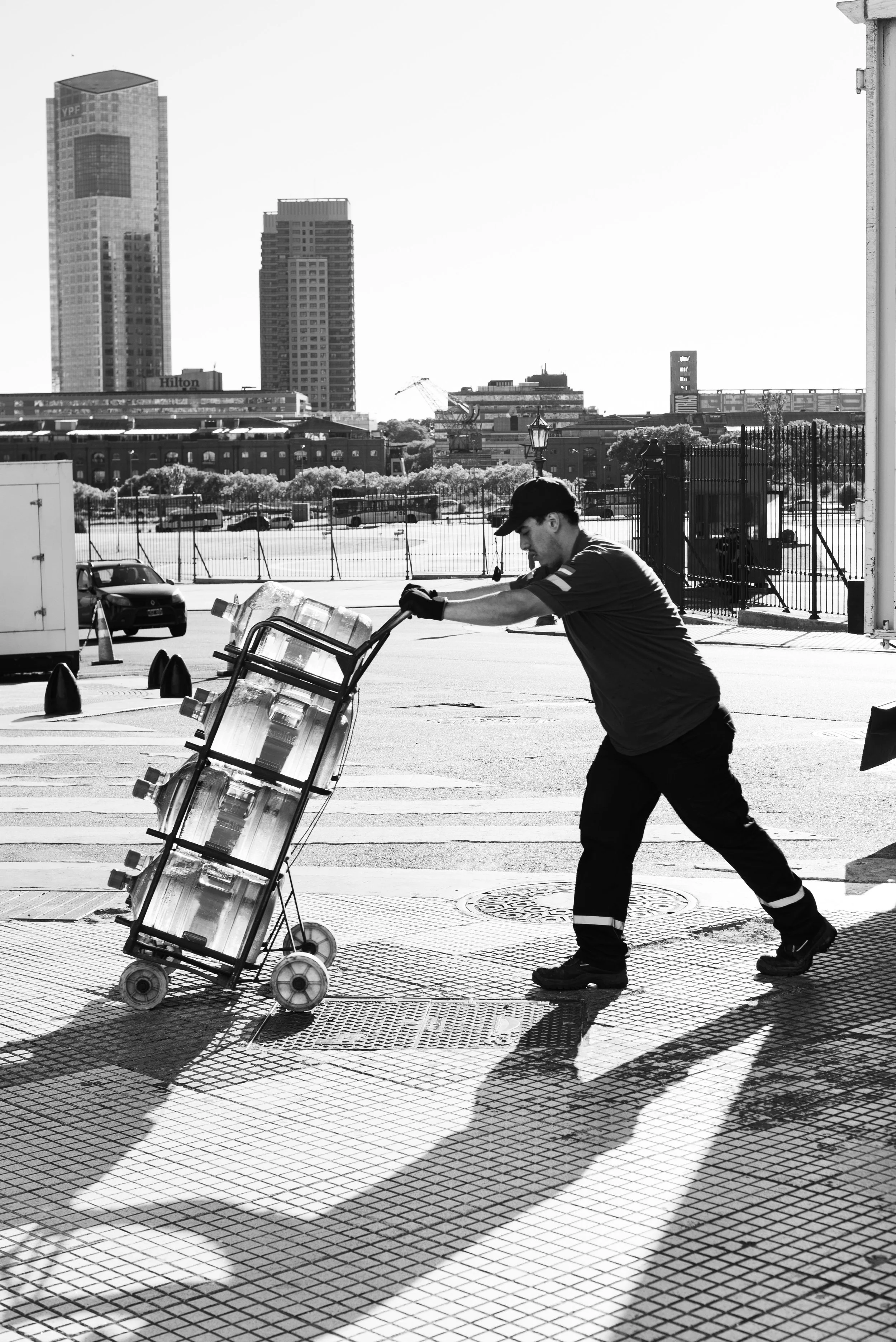 A man dressed in black work clothes and a cap pushing a cart filled with water bottles across a city street, with tall buildings, trees, and parked cars in the background, and shadows cast on the sidewalk.