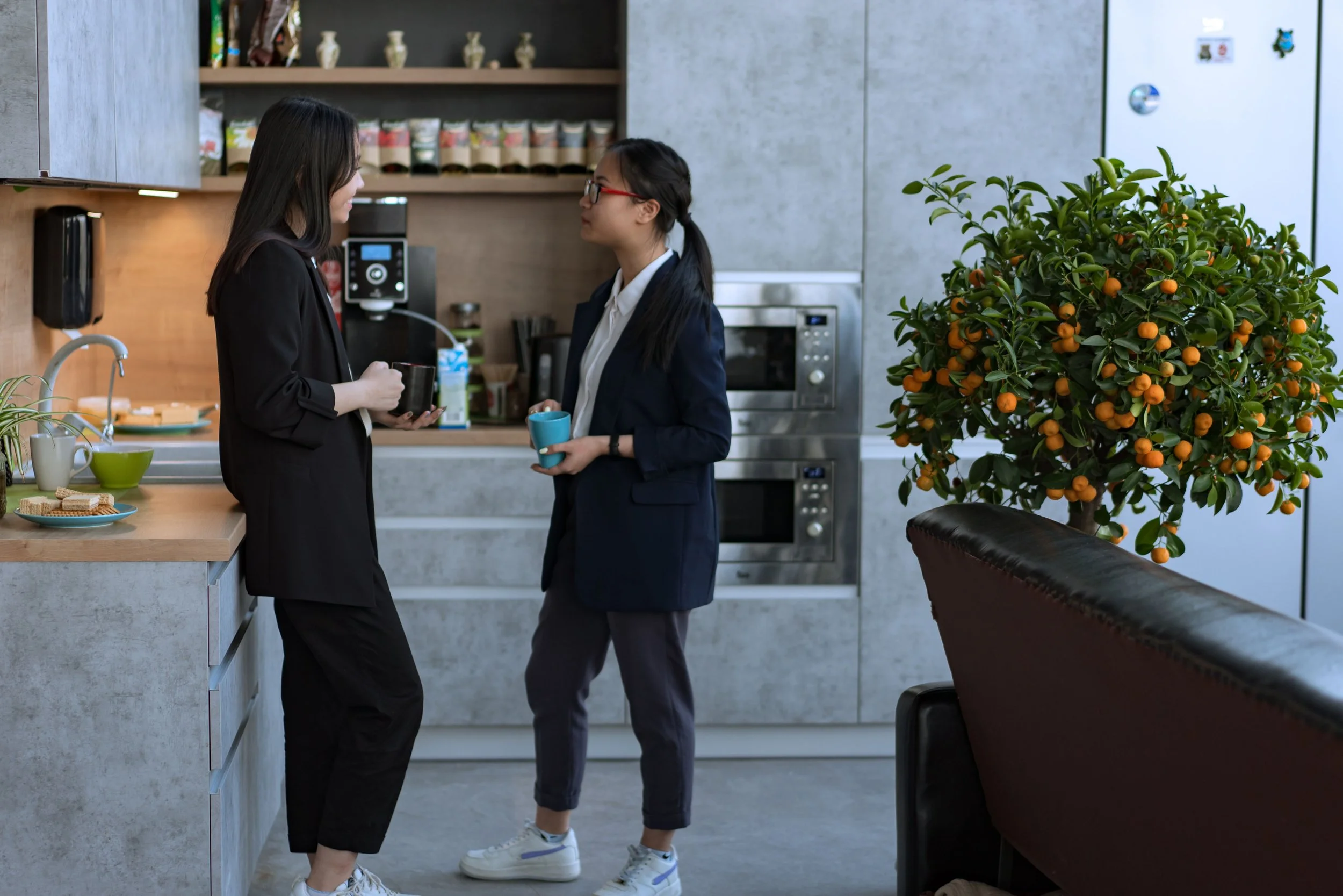 Two women in business attire are having a conversation in a modern office kitchen. One woman is holding a coffee mug, and the other has a blue cup. The kitchen has grey walls, shelves with cups, a microwave, and a large potted plant with orange fruit.