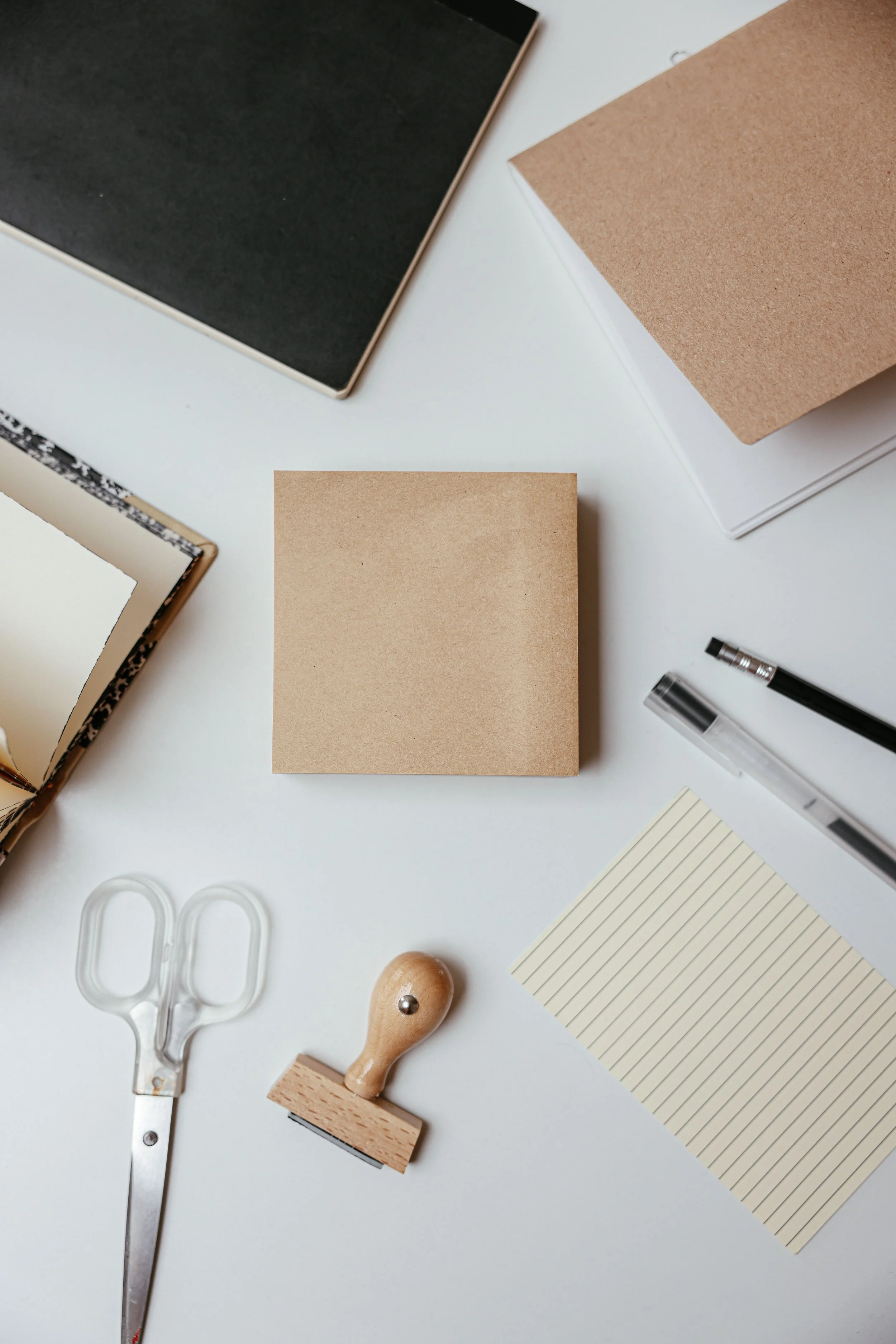Desk with various office supplies including a black-covered notebook, a blank beige sticky note, a black pen, a white pen with a transparent barrel, a pair of scissors, a wooden rubber stamp with a cork handle, a small round wooden object, lined yellow notepad, and a brown paperclip.