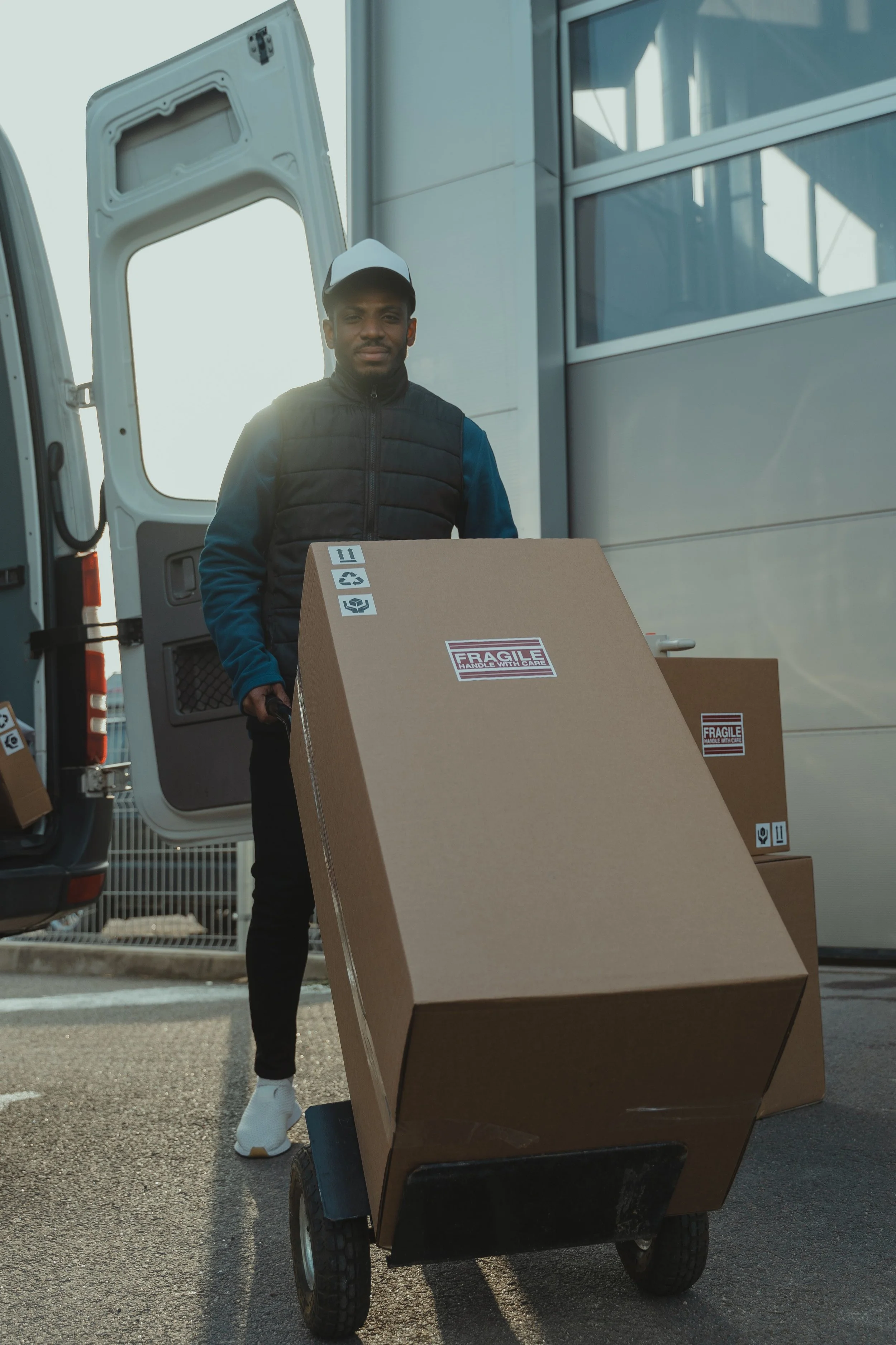A person is standing behind a hand truck loaded with a large cardboard box that has fragile handling labels. The person is wearing a white cap, a dark vest over a blue jacket, and white sneakers. They are near the open back of a delivery van, which is parked outdoors during daylight.