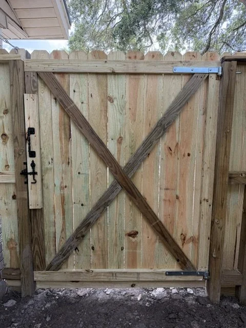 A wooden gate with diagonal and horizontal support beams, metal hinges, and a latch, installed in a backyard.