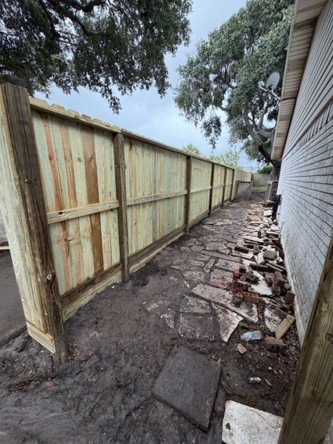 Side yard with a new wooden fence on the left and a stone pathway alongside the house on the right, with scattered bricks and debris.