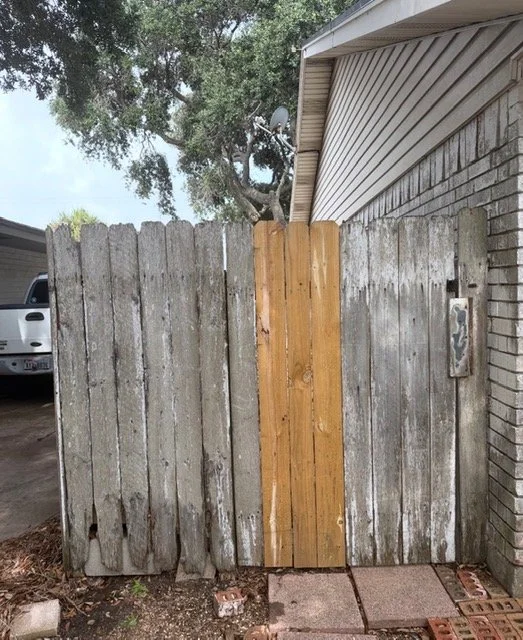 A wooden fence with a new section of lighter wood in the center, attached to a house with brick and siding exterior, and a tree behind it.