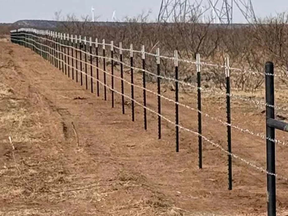 Barbed wire fence running along a barren, brown field with leafless bushes and an overcast sky.