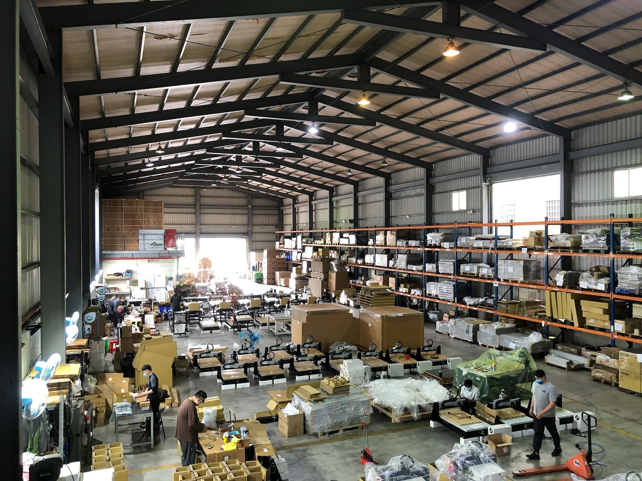 A large warehouse with high ceilings, workers assembling or packaging items at tables, shelving units filled with boxes and supplies, and various equipment used in manufacturing or shipping.