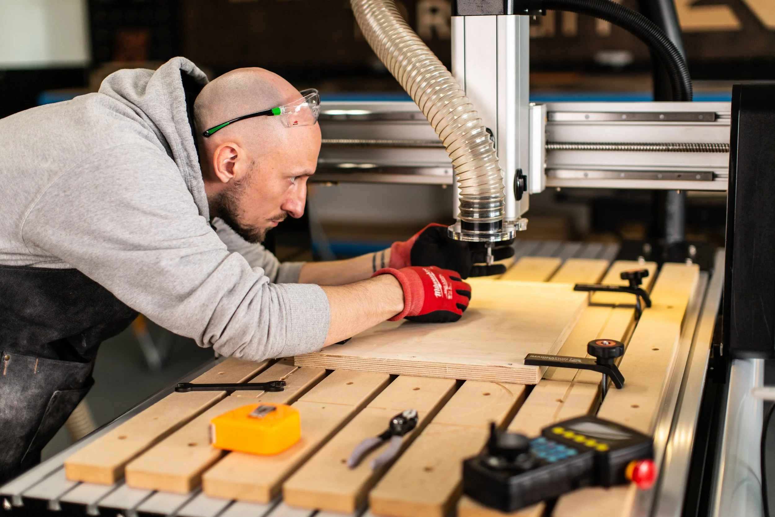 A man wearing safety glasses and red gloves is working on a woodworking project. He is leaning over a piece of wood on a workbench, with a CNC machine or similar woodworking equipment in the background.