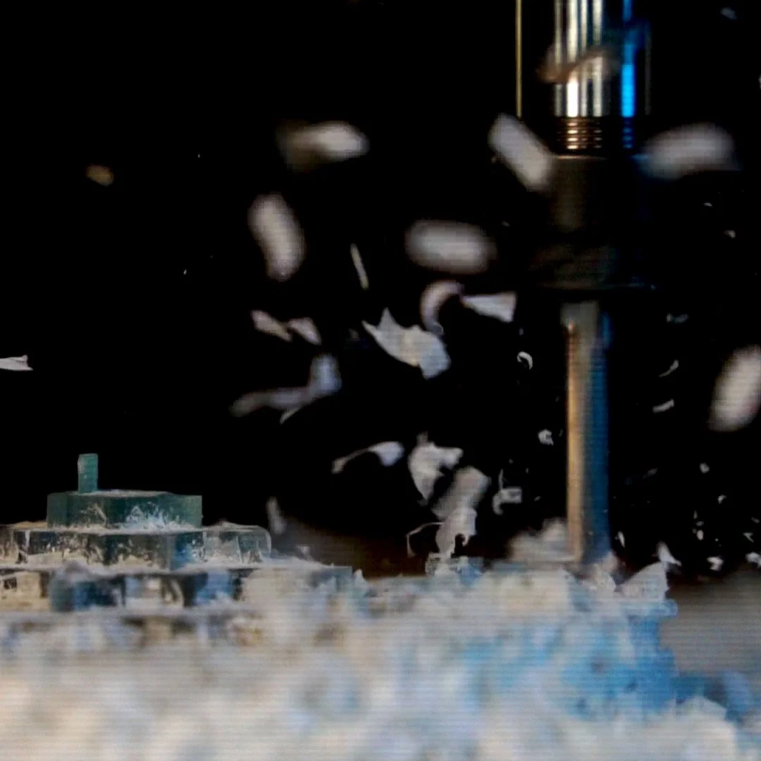 Close-up of small white capsules falling through the air onto a flat surface with machinery in the background.