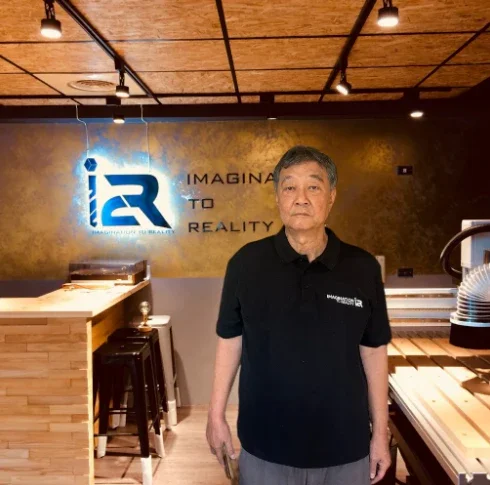 Man in black polo shirt standing inside a modern office or studio with a logo and the words 'IMAGINE TO REALITY' on the wall behind him.