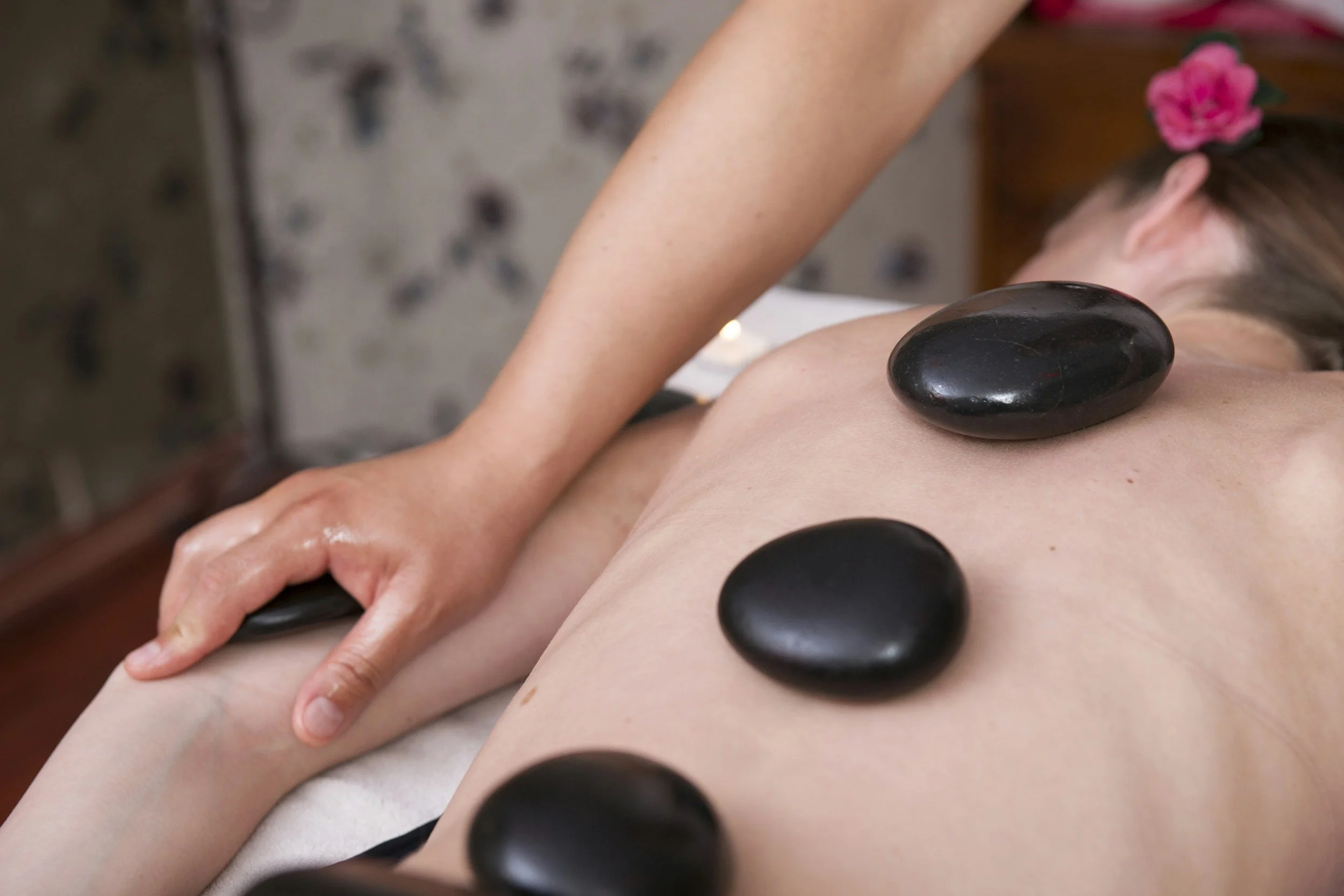A person lying face down receiving a hot stone massage, with black stones placed along their back, in a spa setting with a flower in the background.