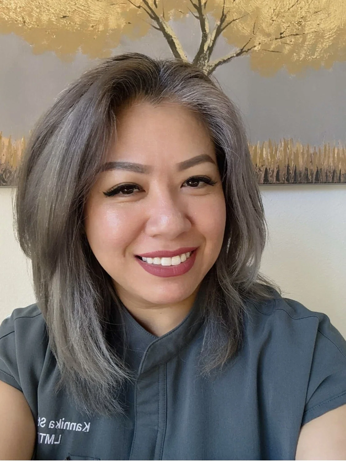 A woman with shoulder-length gray hair and makeup smiling at the camera, wearing a dark gray shirt in front of a painting of a tree with yellow foliage.