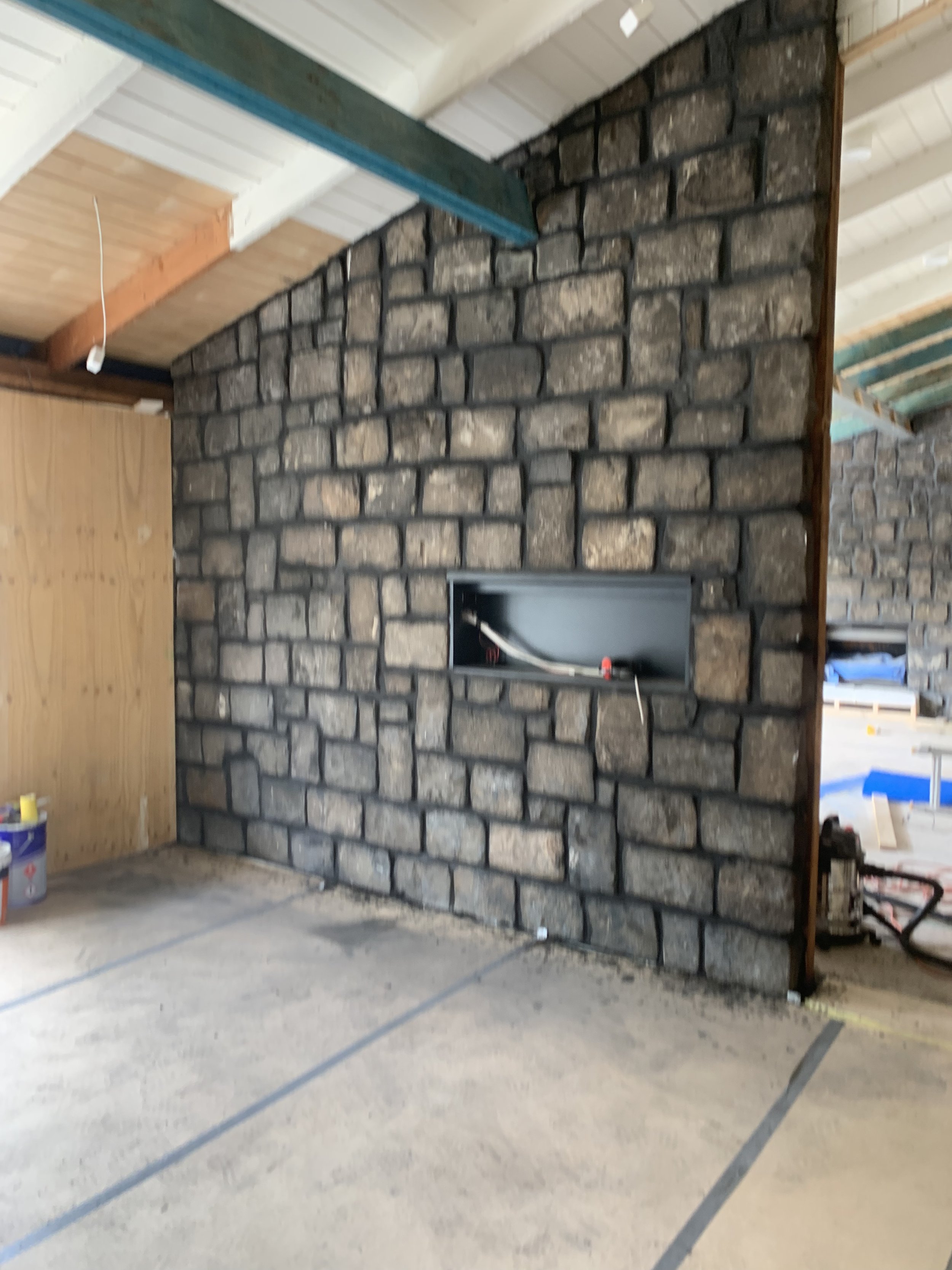 Interior construction scene with a stone fireplace or wall, partially finished, in an unfinished room with wooden ceiling beams and some construction tools and materials scattered around.