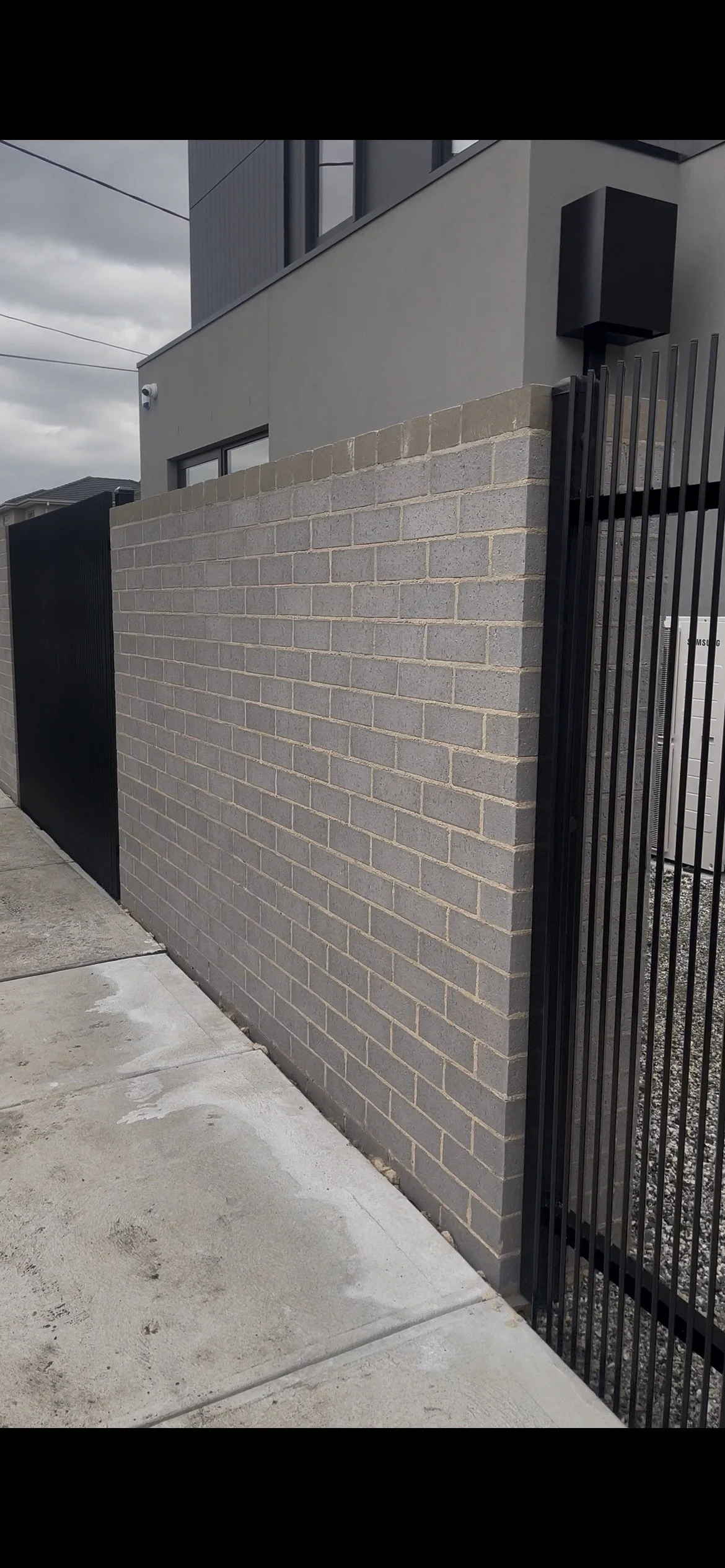 A brick wall and metal gate at the entrance of a modern house with a gray exterior, windows, and an outdoor security camera, under overcast skies.