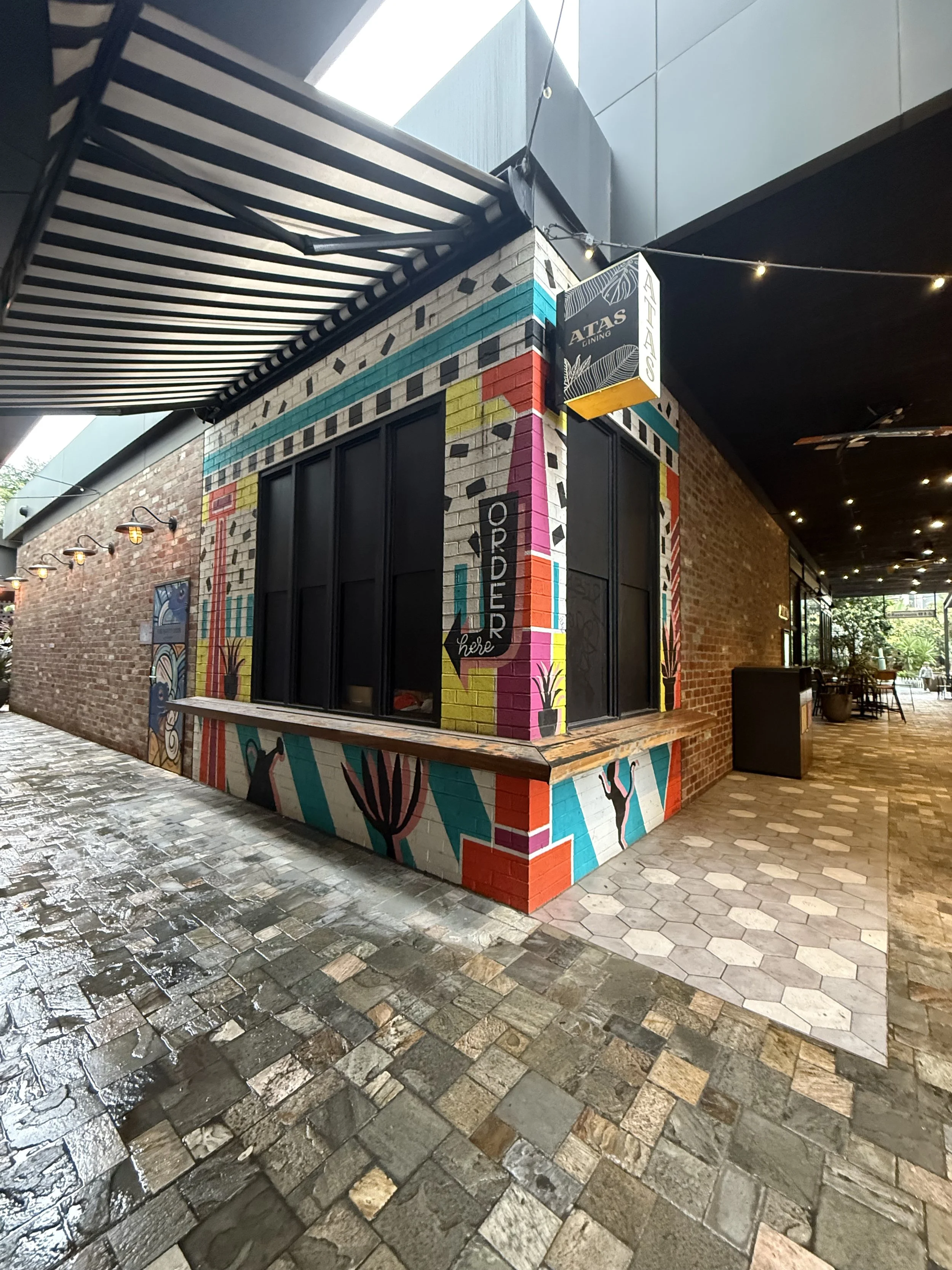 Exterior of a restaurant named 'Atlas Dining' with colorful mural art on brick walls and black window frames. The brick wall is decorated with vibrant geometric and nature-inspired designs. The ground is paved with wet, dark stone tiles, and there are small outdoor tables and chairs in the background, with string lights above.