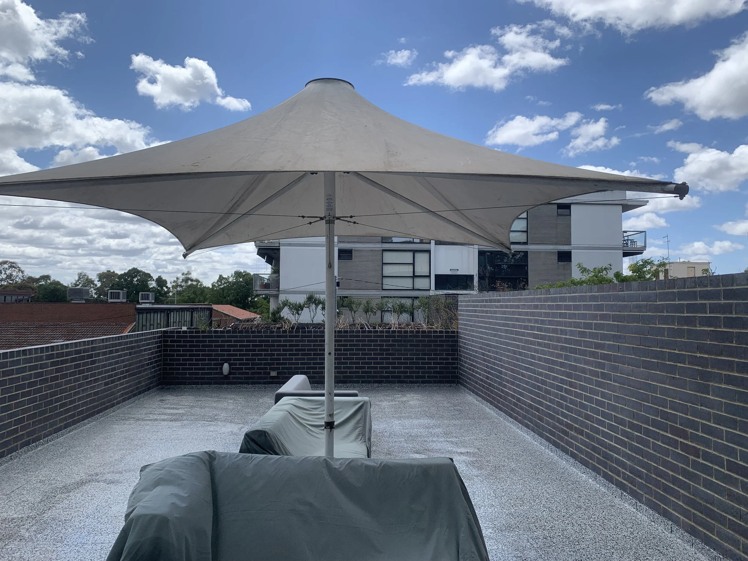 View of a rooftop patio with a large white outdoor umbrella, covered furniture, a brick wall, and neighboring buildings under a partly cloudy sky.
