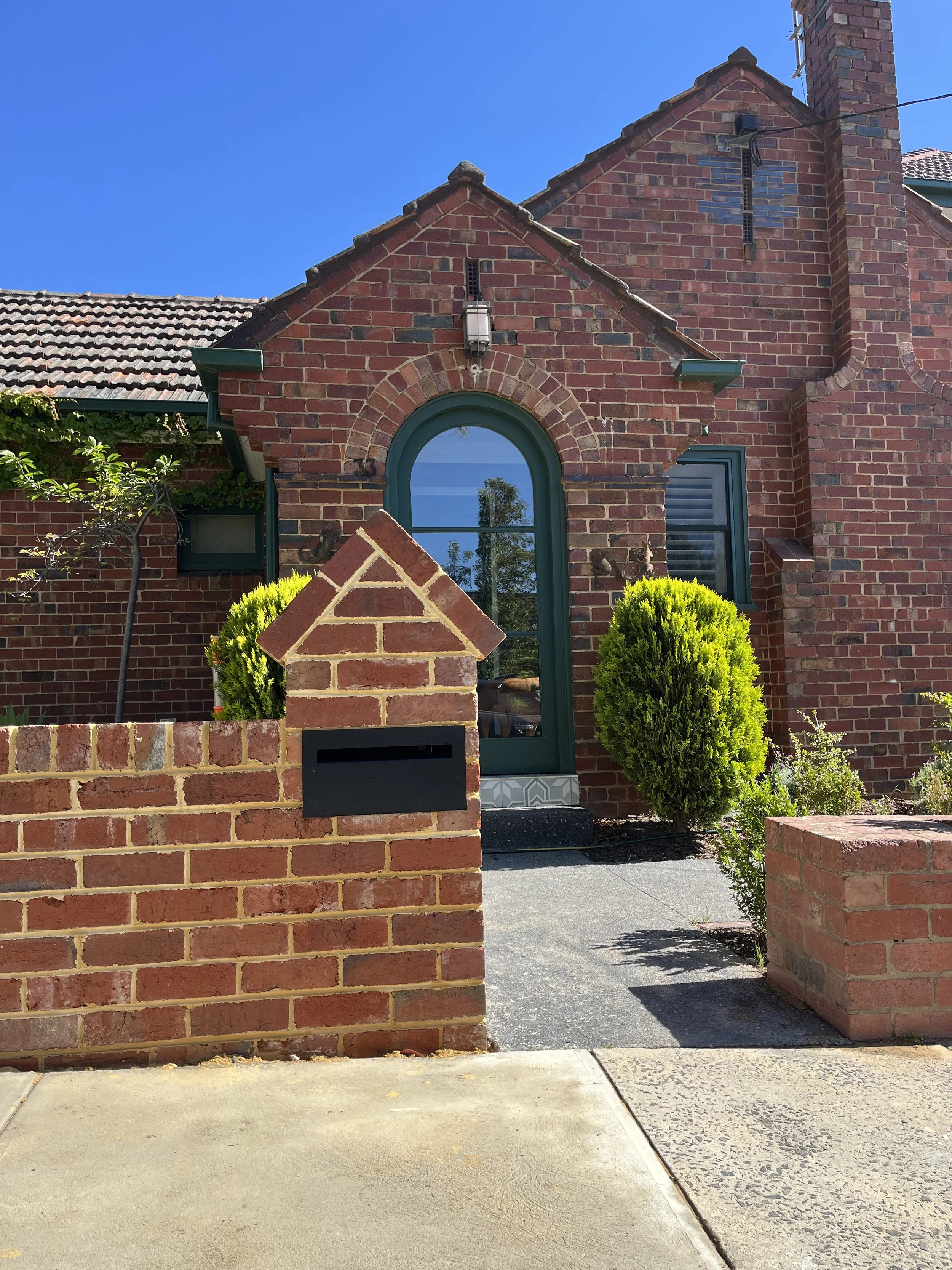 Front view of a red brick house with arched window, small shrubs, brick fence with mailbox, and a sidewalk in the foreground under a blue sky.