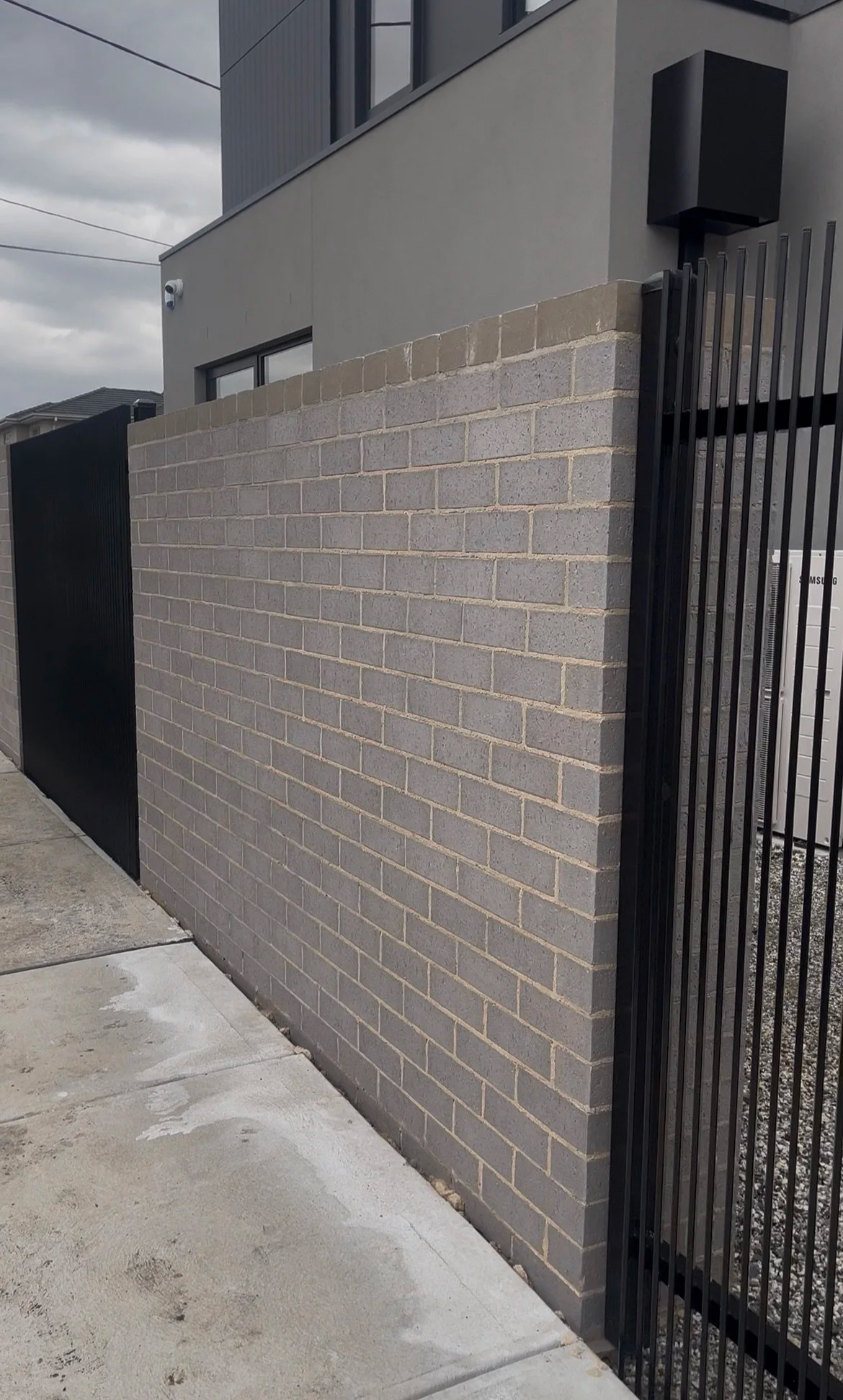 Photo of a modern building with gray exterior walls, a brick wall, a security camera, and a black metal gate with vertical bars.